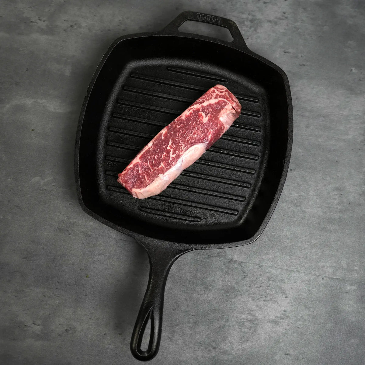 Prime Holstein New York Strip Steak, a well-marbled end cut, sits in a square cast-iron grill pan, showcasing its rich texture and quality.