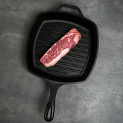 Prime Holstein New York Strip Steak, a well-marbled end cut, sits in a square cast-iron grill pan, showcasing its rich texture and quality.