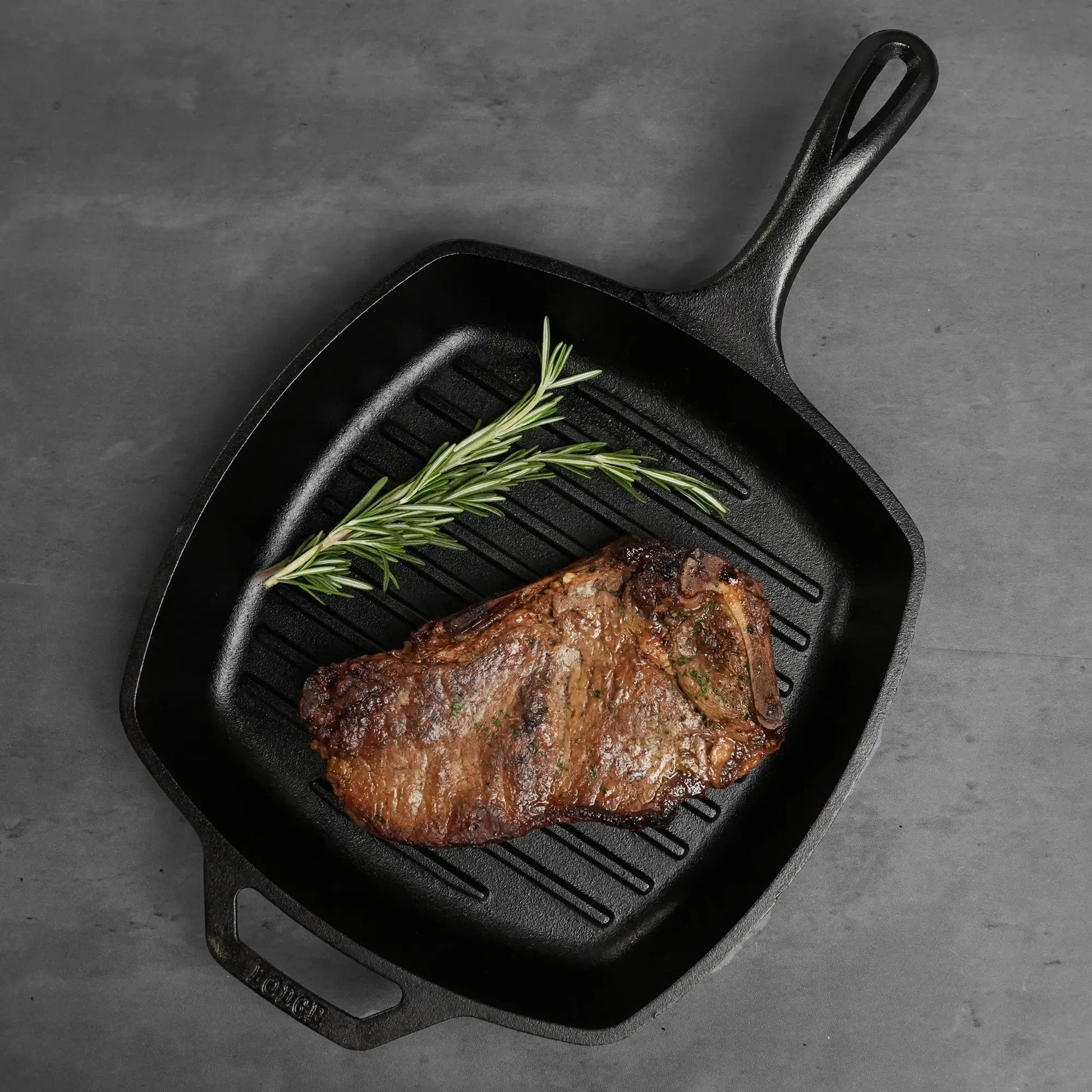 Square cast-iron grill pan with a seared Dry Aged Prime Bone In Kansas City Strip Steak, accompanied by a fresh rosemary sprig, highlighting rich marbling and dry-aged perfection.