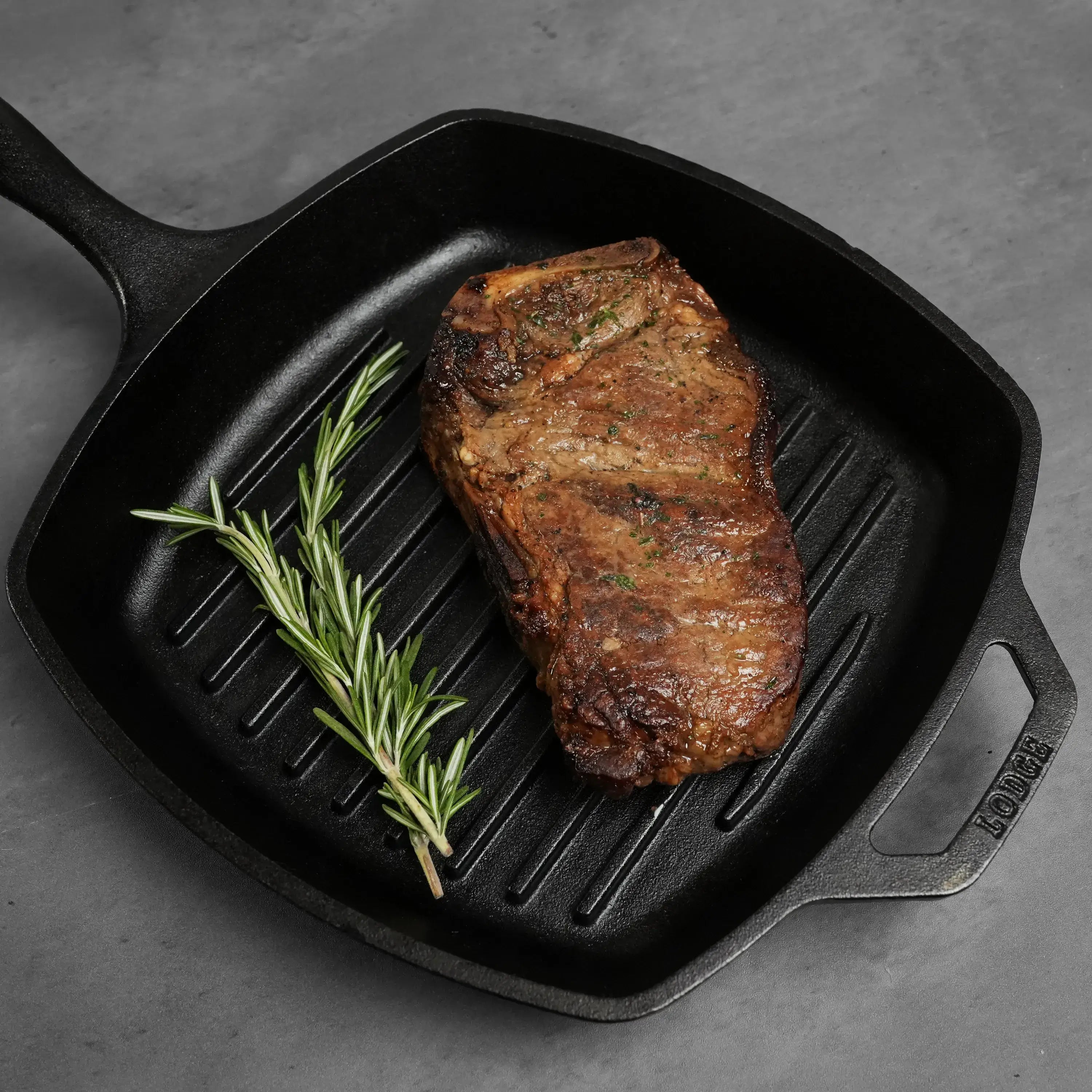 Seared Dry Aged Prime Bone In Kansas City Strip Steak in a cast-iron grill pan, garnished with fresh rosemary, highlighting its juicy, marbled texture.