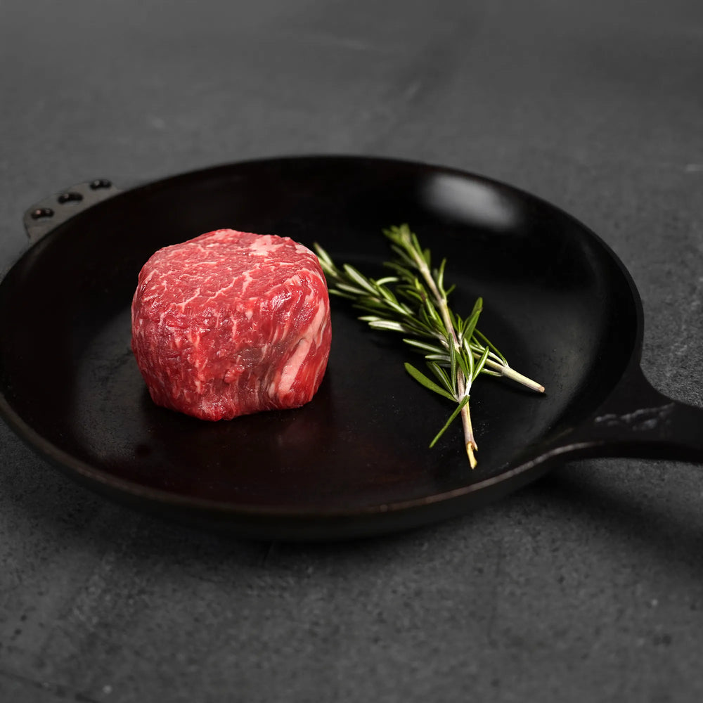 Raw steak on black iron pan with herbs on dark surface.