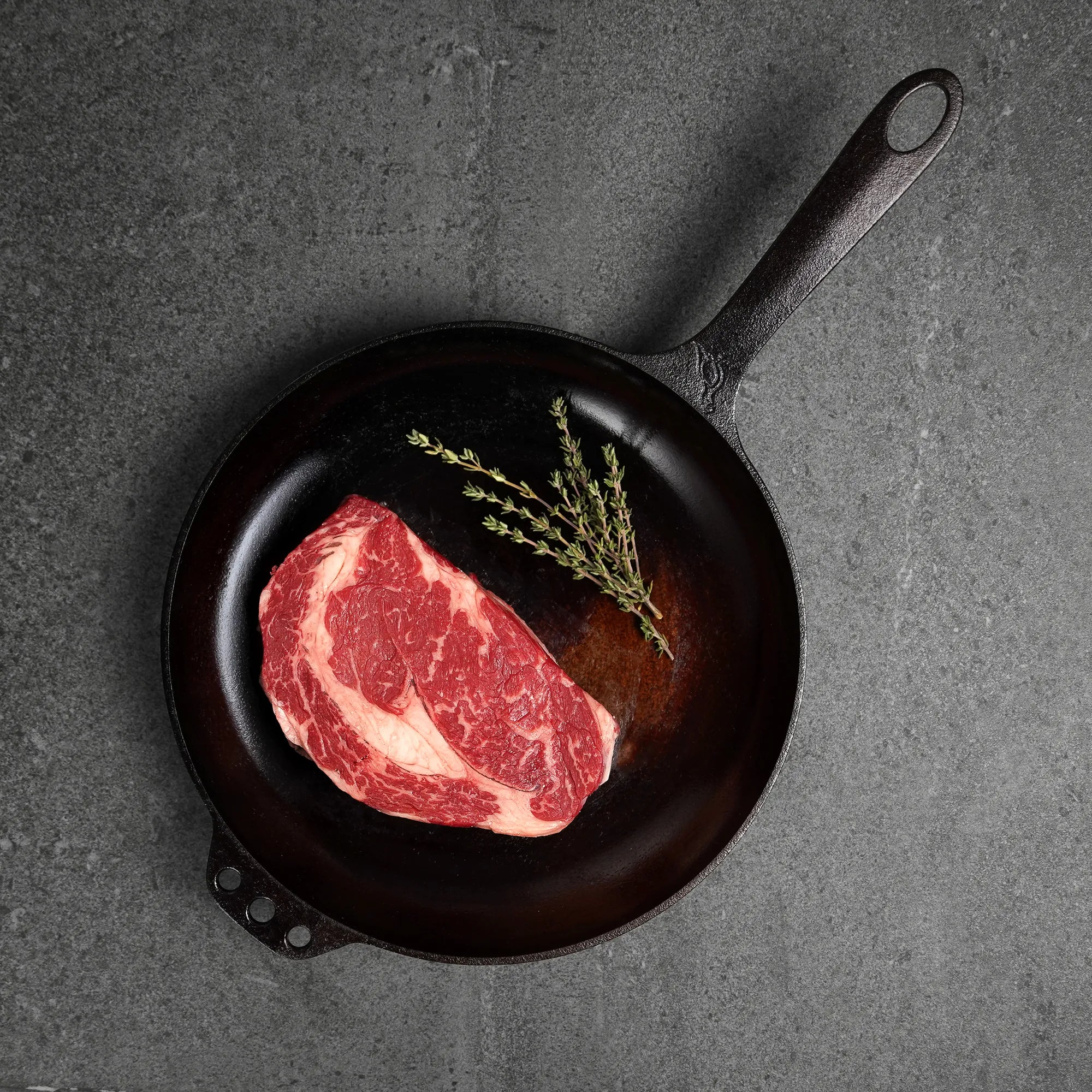 Raw steak on a black cast iron skillet with a sprig of rosemary on a dark gray background