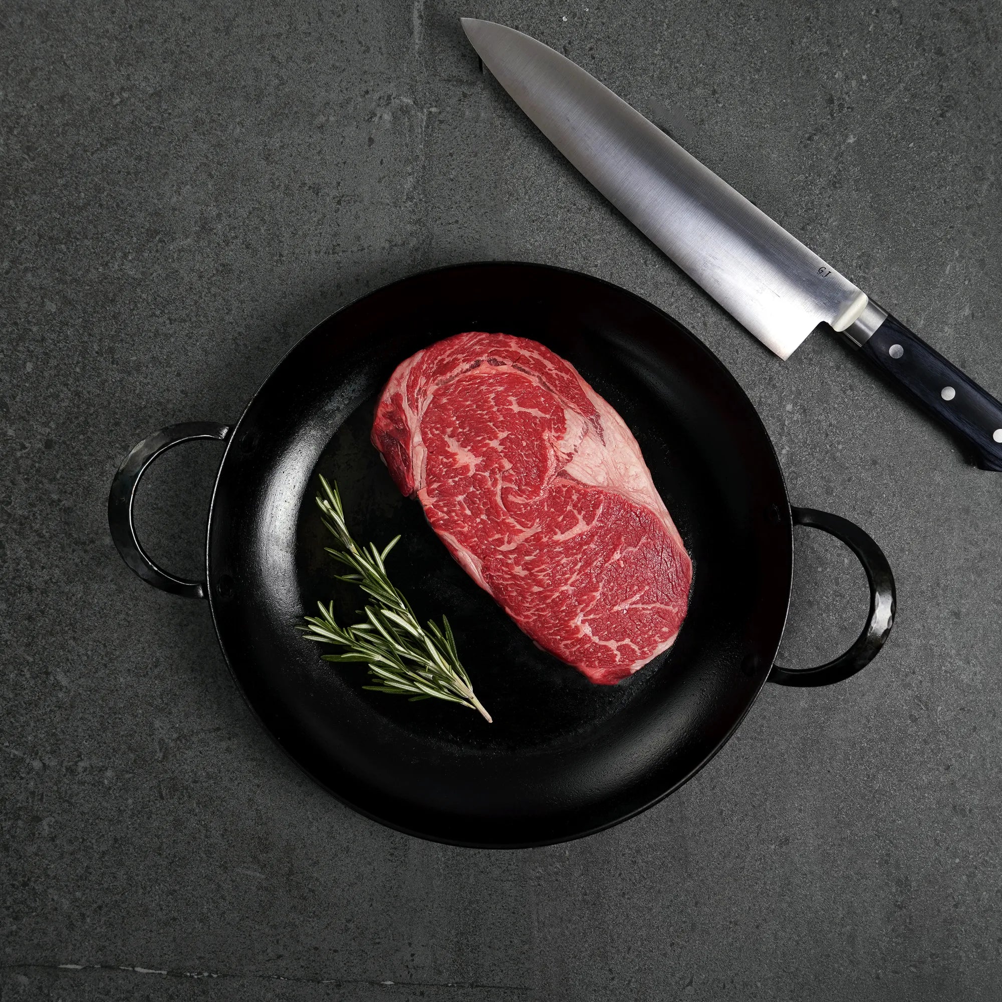 Raw steak on black iron pan with rosemary garnish and silver knife