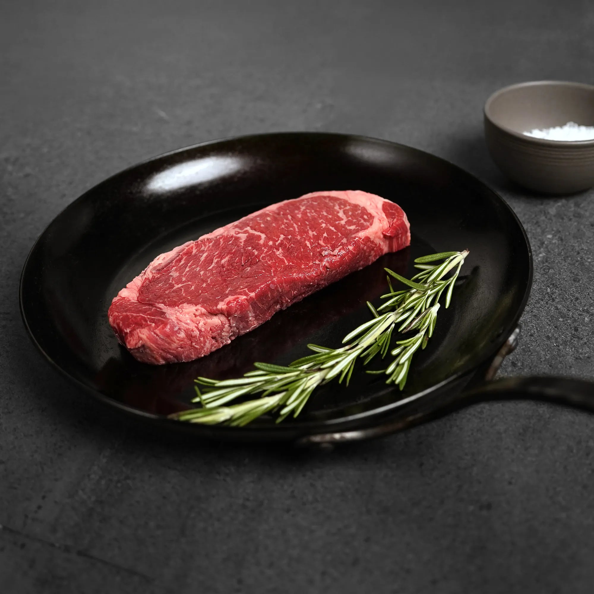 Raw steak on a black pan with rosemary and salt in the background