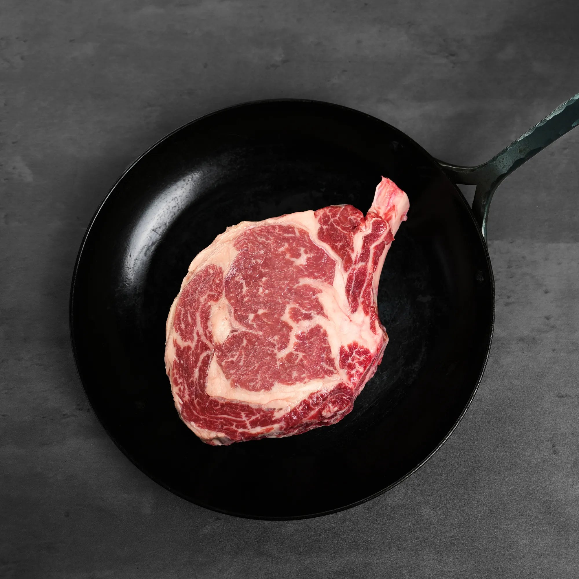 Prime Bone In Cowboy Ribeye Steak in skillet; well-marbled, thick cut for even cooking, enhancing flavor for grilling, pan-searing, or broiling.