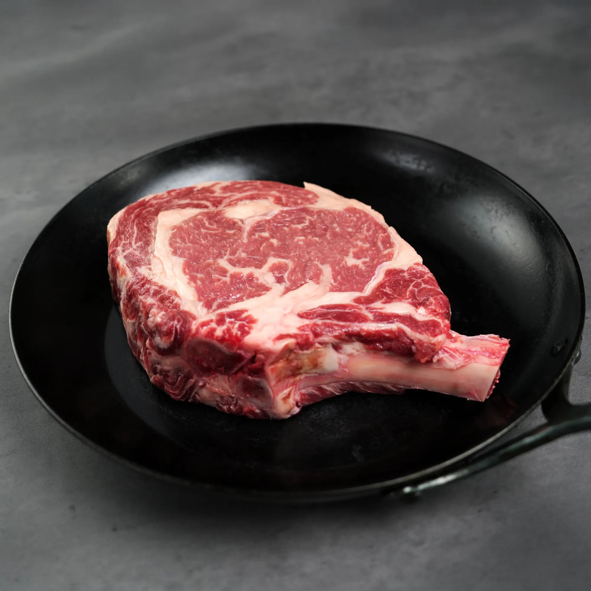 Prime Bone In Cowboy Ribeye Steak in a black cast-iron pan, showcasing its precise cut and thickness for enhanced flavor and even cooking.
