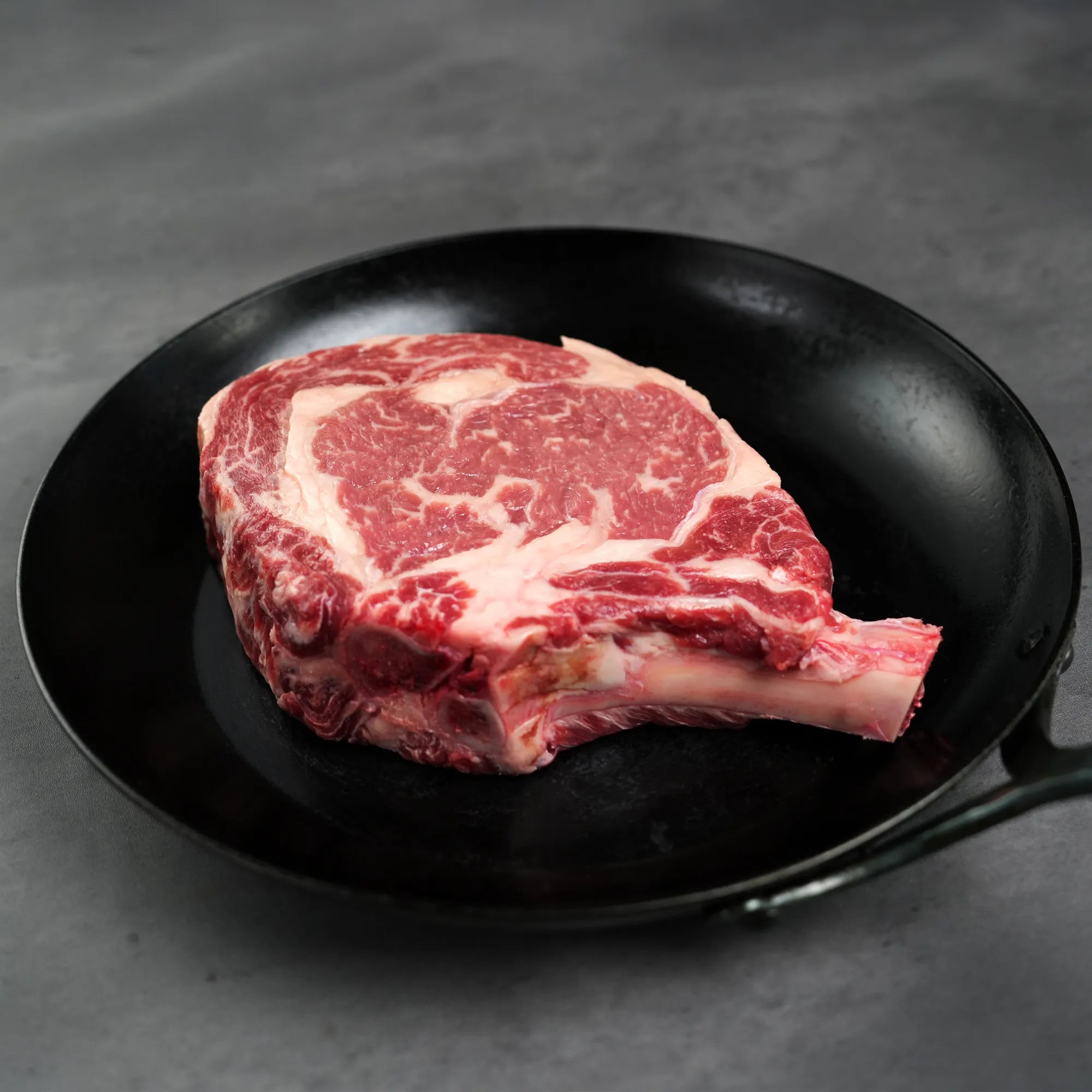 Prime Bone In Cowboy Ribeye Steak in a black cast-iron pan, showcasing its precise cut and thickness for enhanced flavor and even cooking.