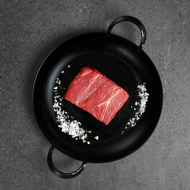 Raw steak on a bed of salt in a black frying pan on a dark surface