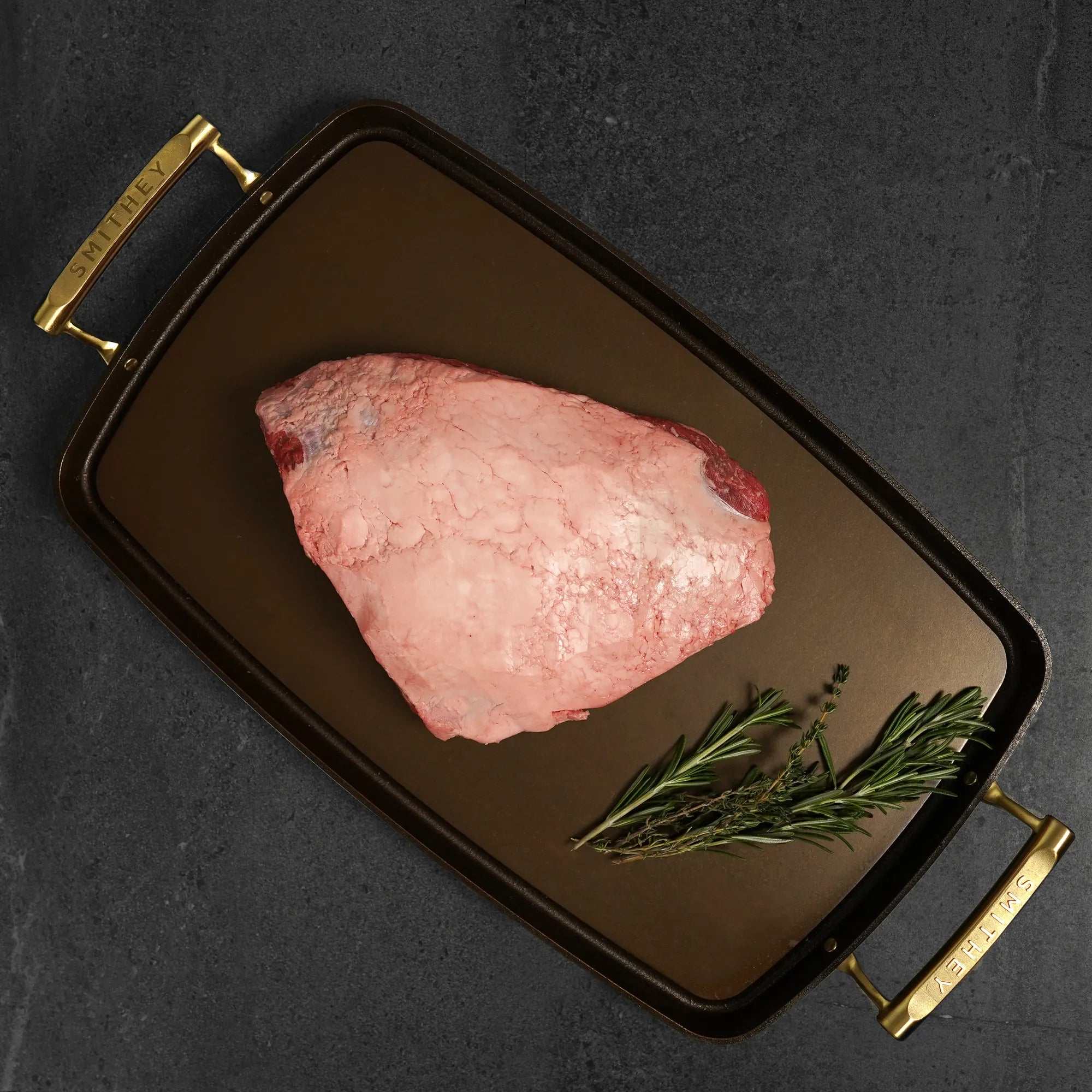 Raw meat on a dark serving tray with gold handles and rosemary sprigs on a dark background