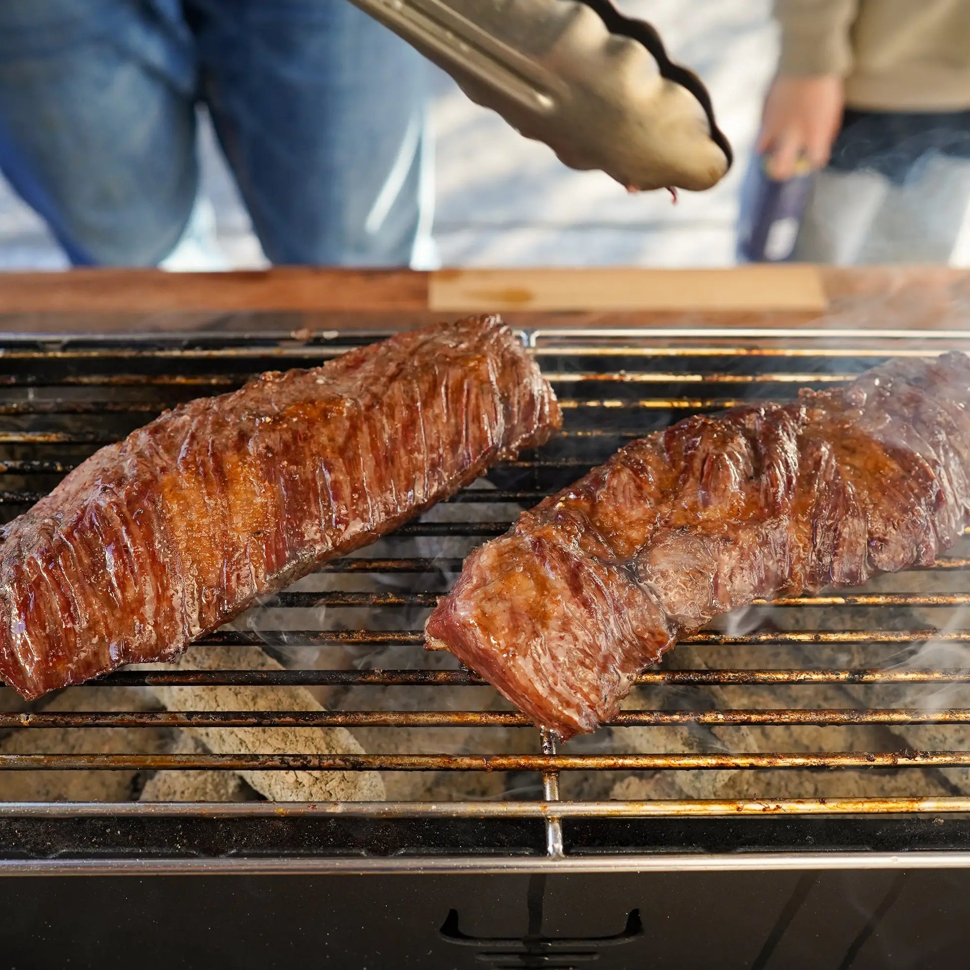 Prime Skirt Steak sizzling on a charcoal grill, with glowing coals beneath and a person using tongs to turn the flavorful beef cut.