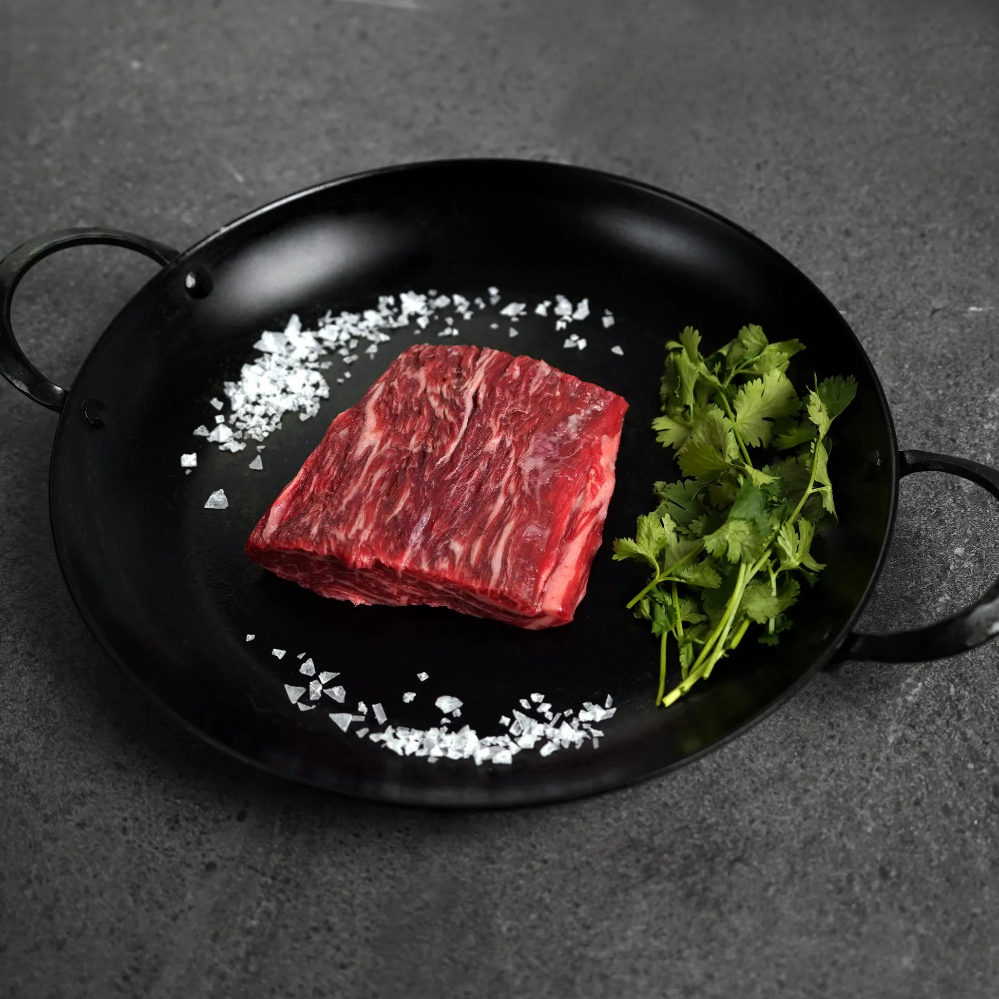 Raw beef on a black pan with salt and herbs on a dark surface