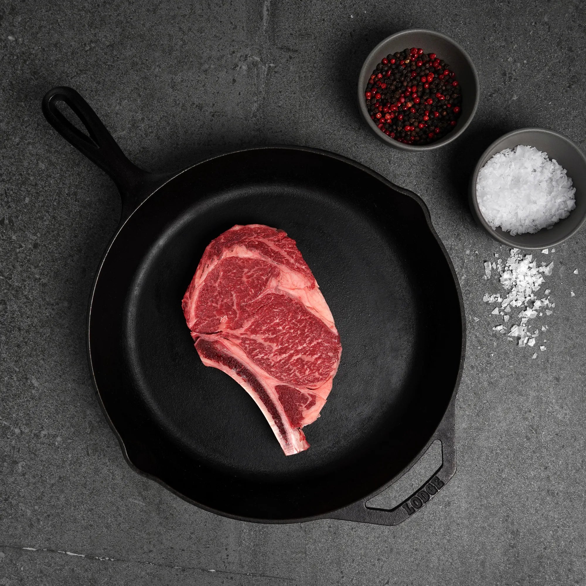 Raw steak on a black cast iron skillet with salt and pepper bowls on a dark surface