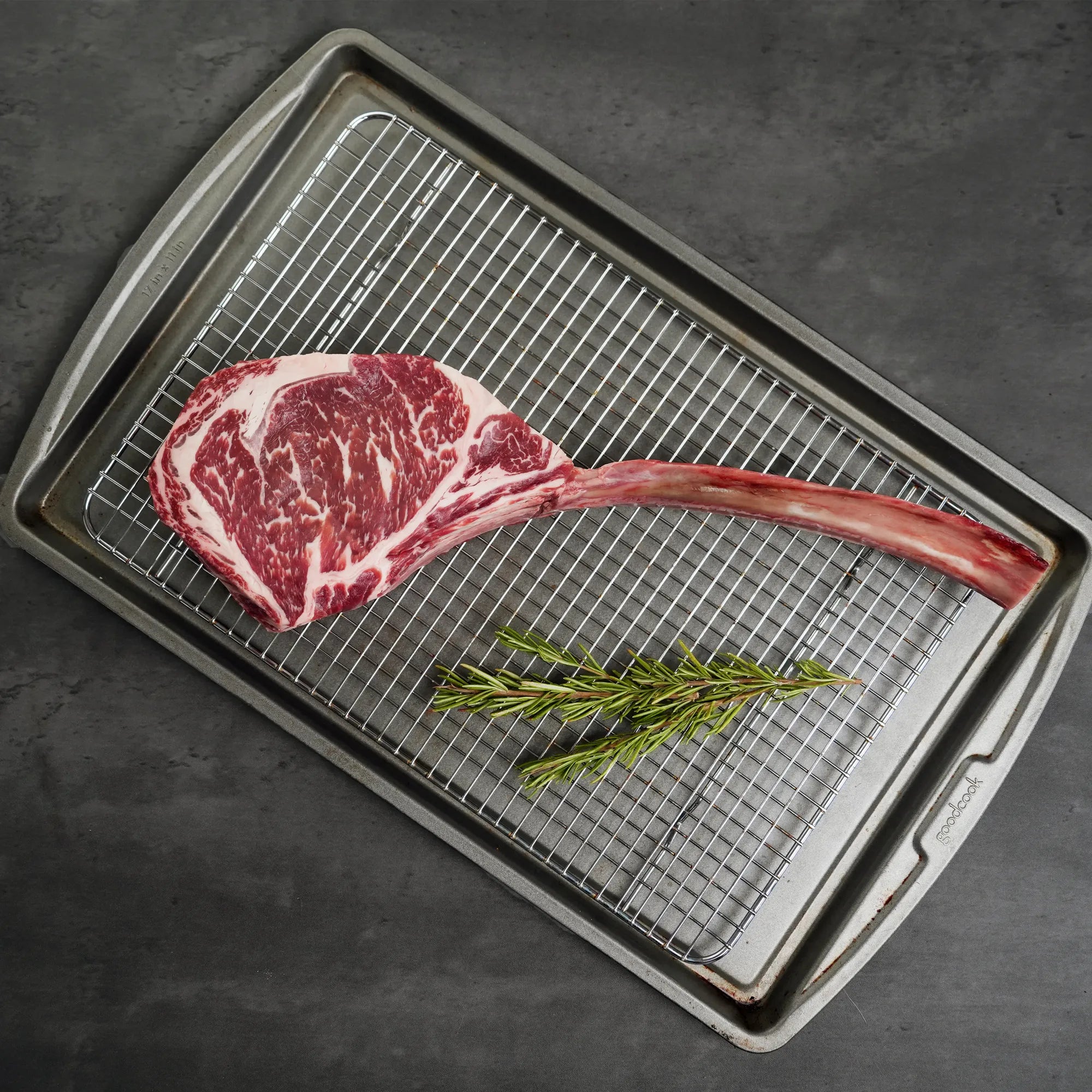 Raw ribeye steak on a metal grill pan with rosemary on a dark surface