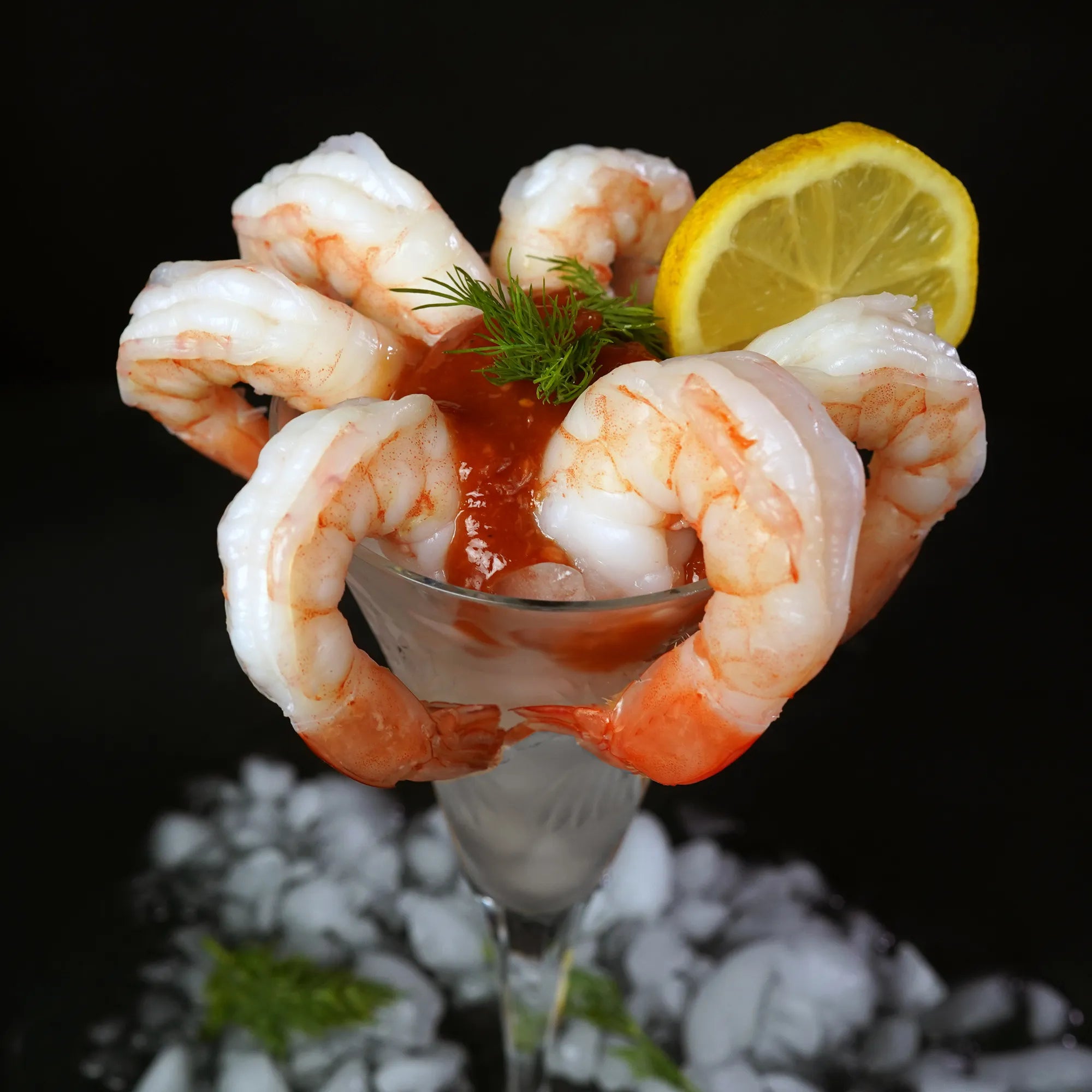 Jumbo Cooked Shrimp elegantly arranged around a cocktail glass with sauce, lemon wedge, and dill garnish, set on crushed ice, perfect for easy serving.