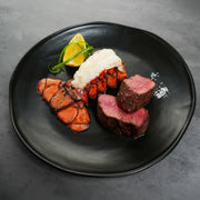 Surf & Turf XL Combo: A lobster tail with elevated meat, medium-rare filet mignon medallions, lemon wedge, herbs, and coarse salt on a black plate.