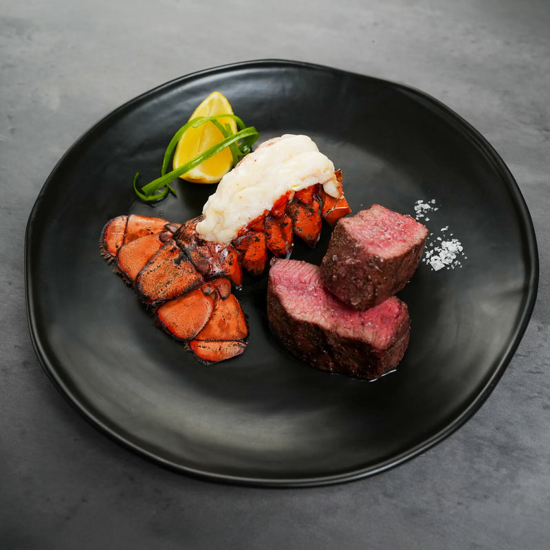 Surf & Turf Combo: Grilled lobster tail and medium-rare beef tenderloin slices, garnished with lemon, herbs, and coarse salt on a black plate.