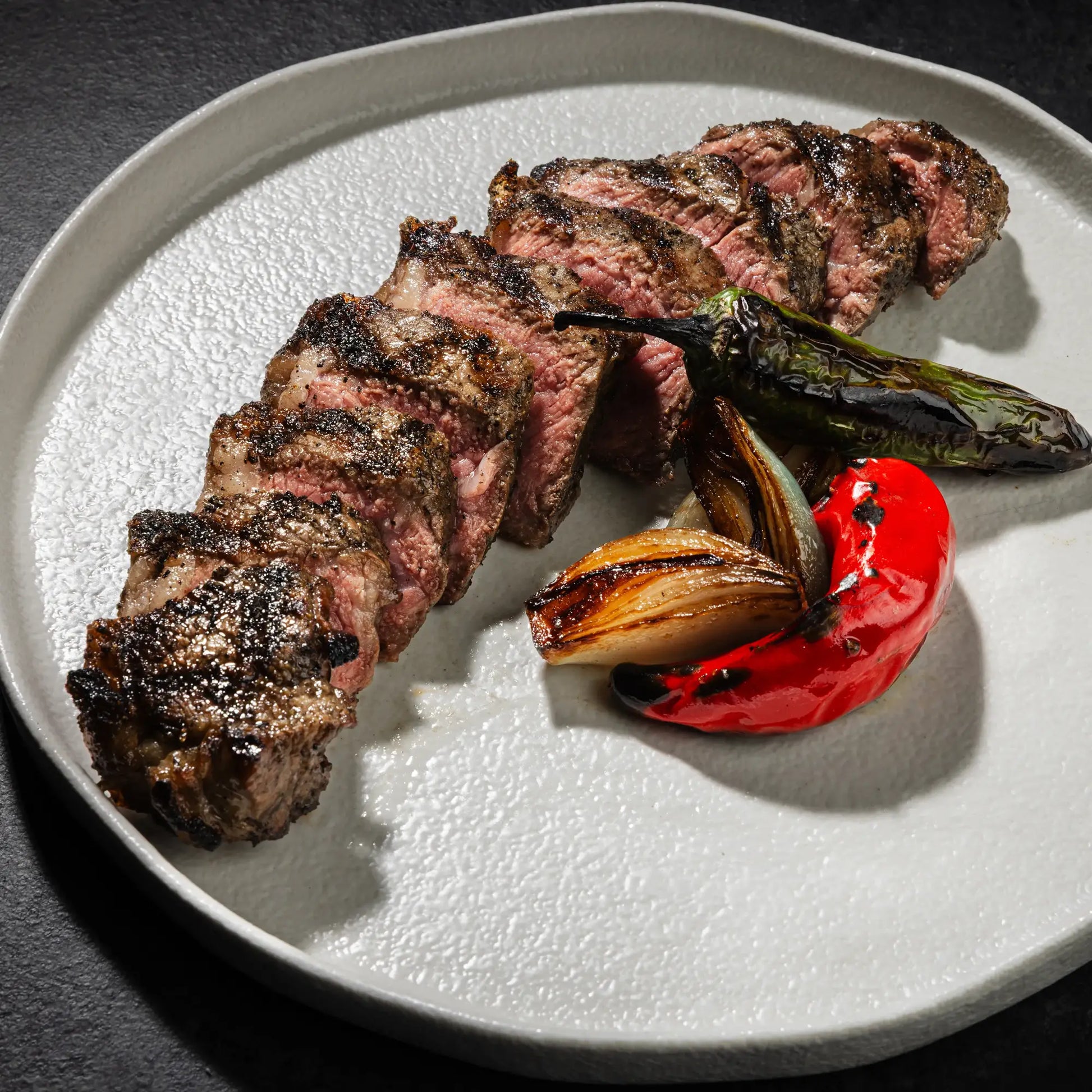 Dry Aged Prime New York Strip Steak Petite, sliced medium-rare, with char-grilled peppers and onions on a ceramic plate, showcasing its tender, beefy flavor.