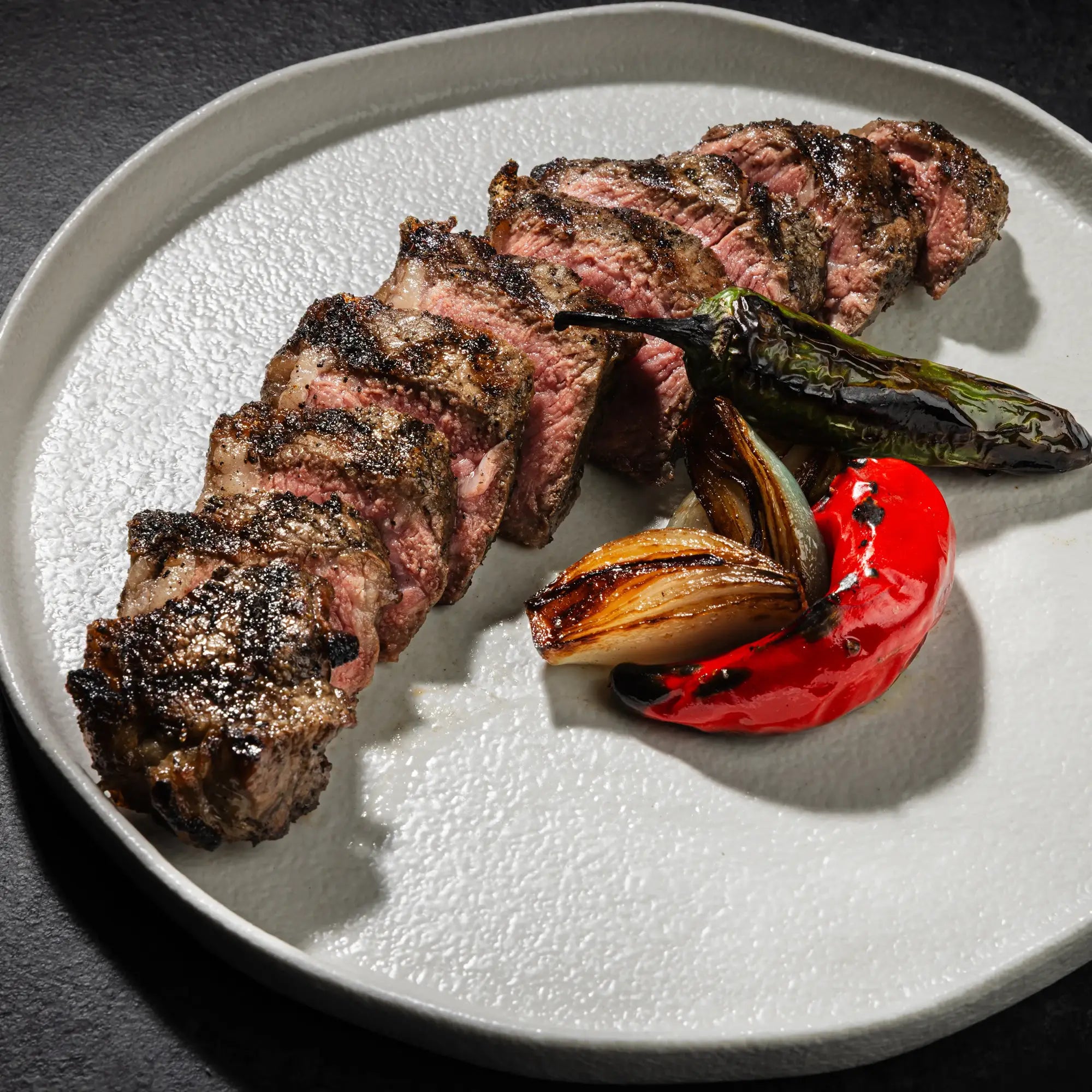 Dry Aged Prime New York Strip Steak Petite, sliced medium-rare, with char-grilled peppers and onions on a ceramic plate, showcasing its tender, beefy flavor.