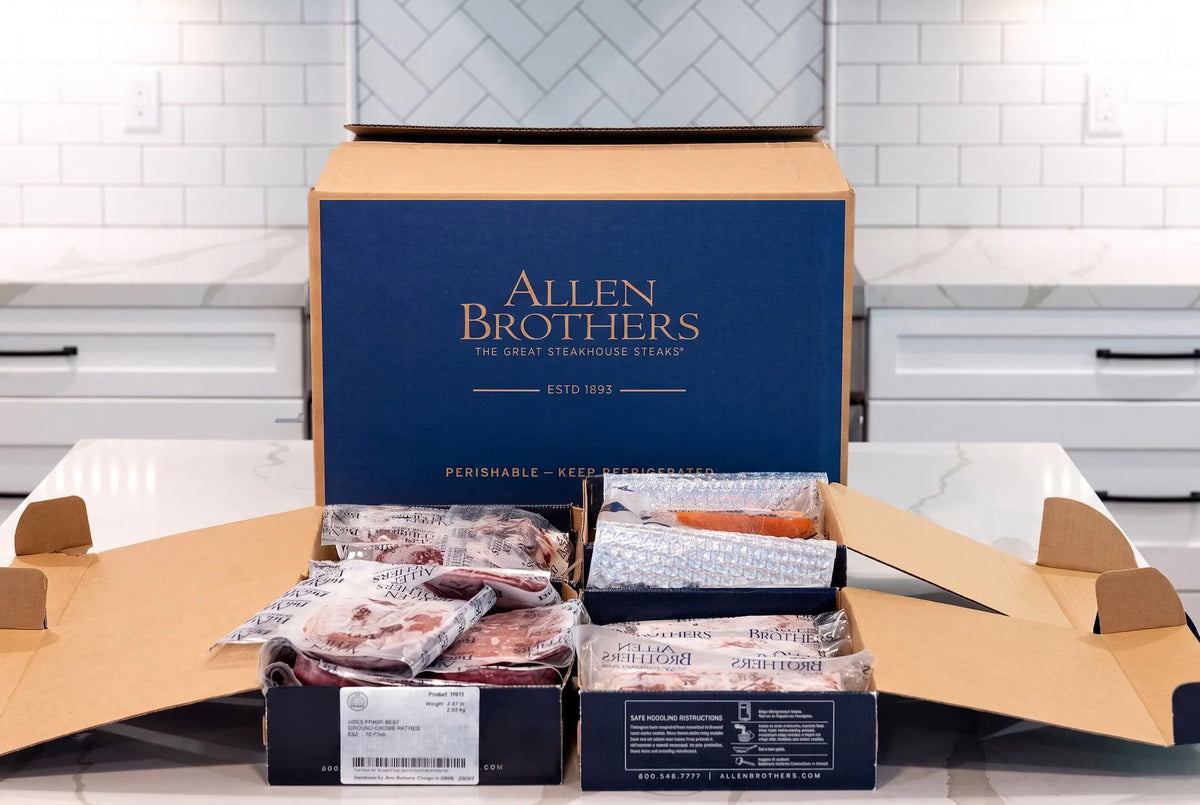 An open Allen Brothers meat delivery box displayed on a kitchen counter, with the branded navy blue box standing behind it and packaged meats visible inside.