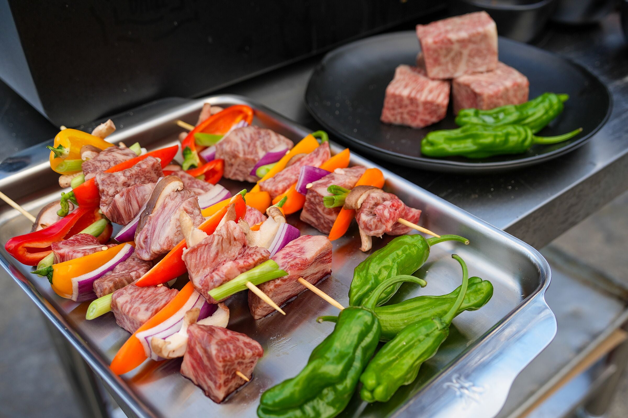 Skewers of meat and vegetables on a metal tray with a side of raw meat and green peppers on a black plate.