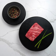 Rosewood Texas Wagyu Bavette Steak, highly marbled, on a black plate with chive stems, adjacent to a bowl of mixed peppercorns on a marble surface.