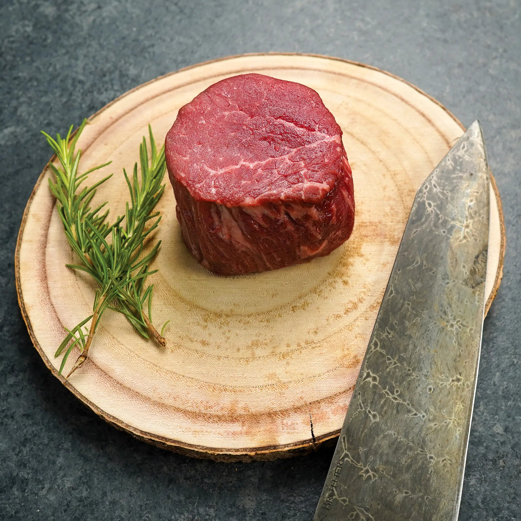 Surf & Turf XL Combo: A raw beef filet mignon on a wooden board with rosemary and a patterned knife, highlighting premium quality and culinary readiness.