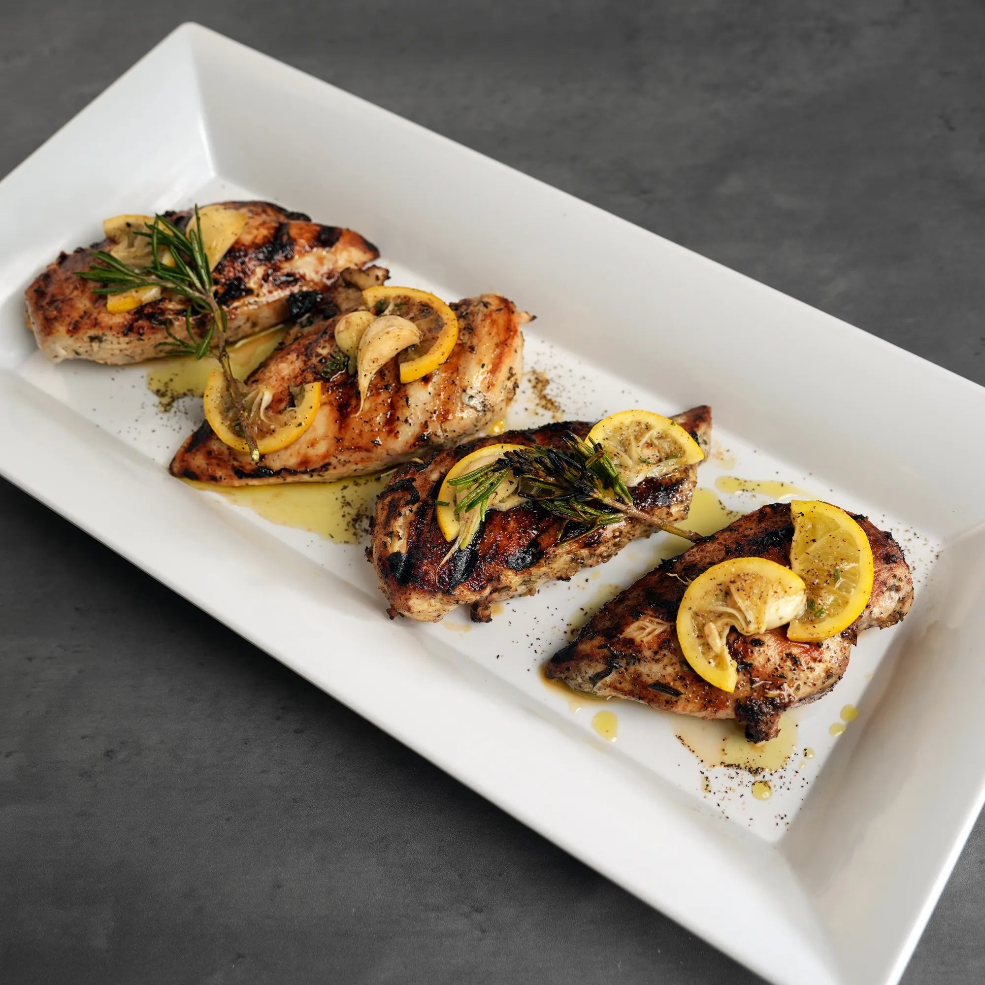 All-Natural Chicken Breasts: Grilled, char-marked pieces garnished with oil, cracked pepper, herbs, lemon slices, garlic cloves, and rosemary. Raised in the USA, no additives.