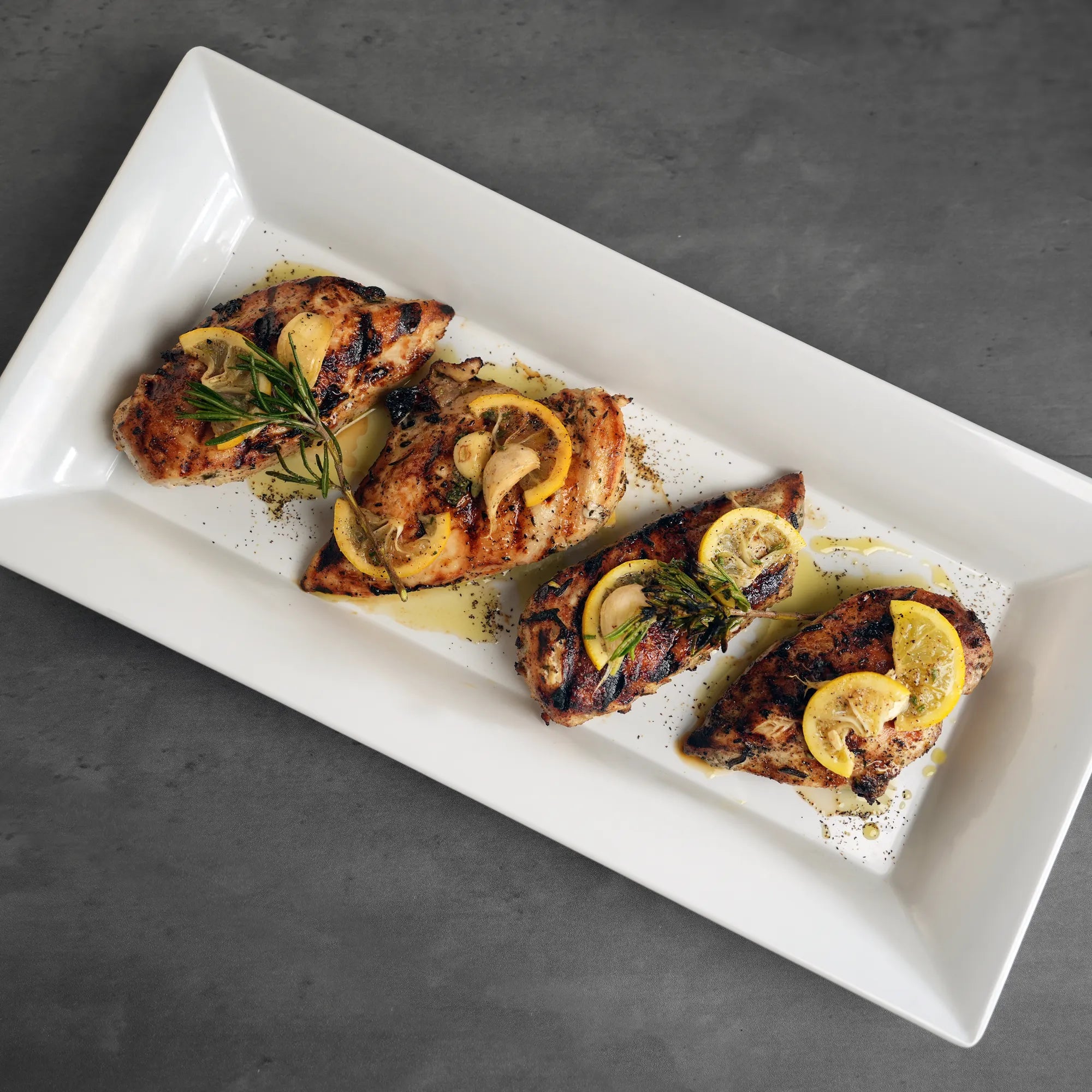 All-Natural Chicken Breasts on a white platter, grilled with lemon slices, garlic, rosemary, showcasing high-quality, hormone-free U.S. raised chicken, ready to ship frozen.