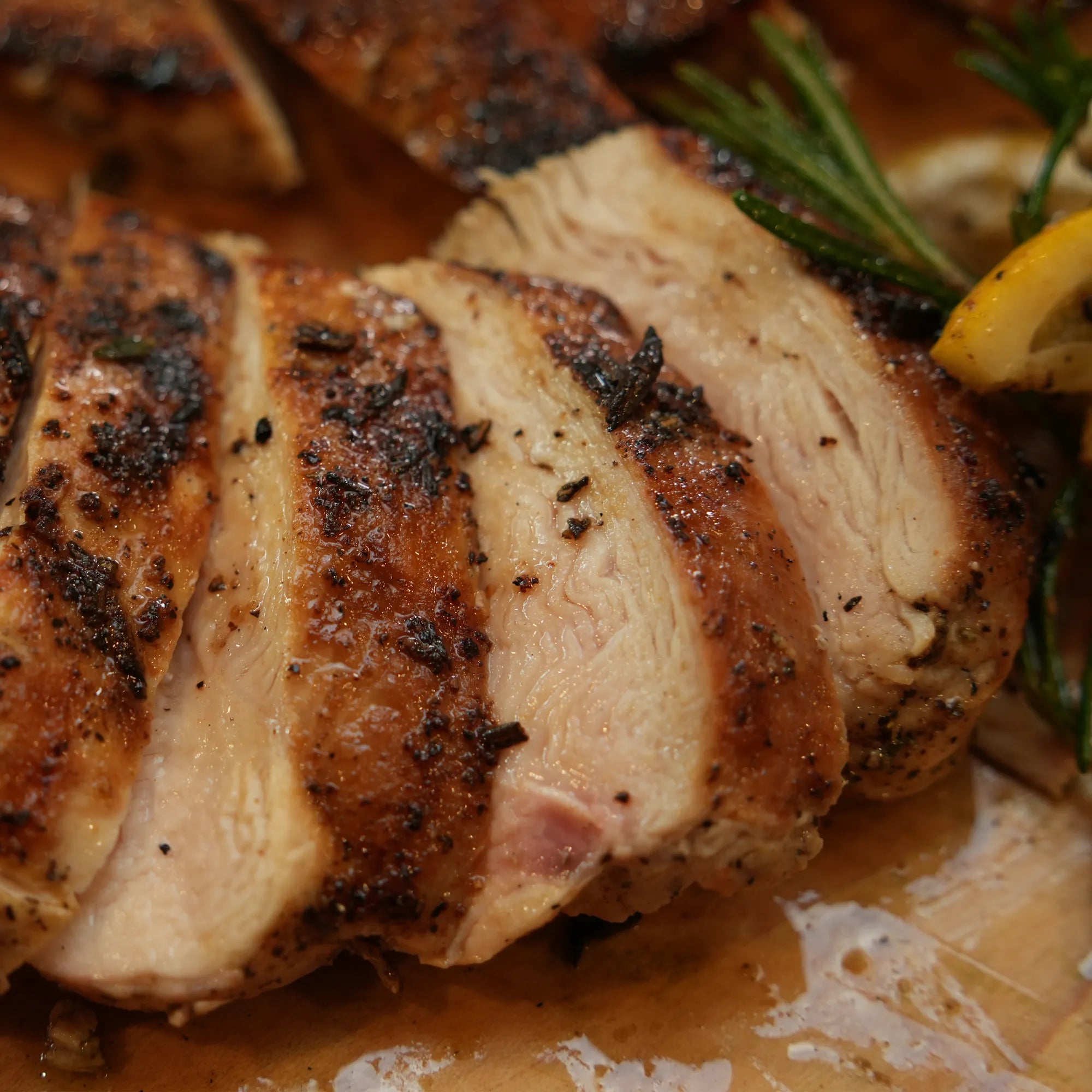 Pasture Raised Heritage Split Chicken, thinly sliced and seasoned with black pepper and rosemary, on a cutting board with lemon and herb garnish, reflecting its juices.