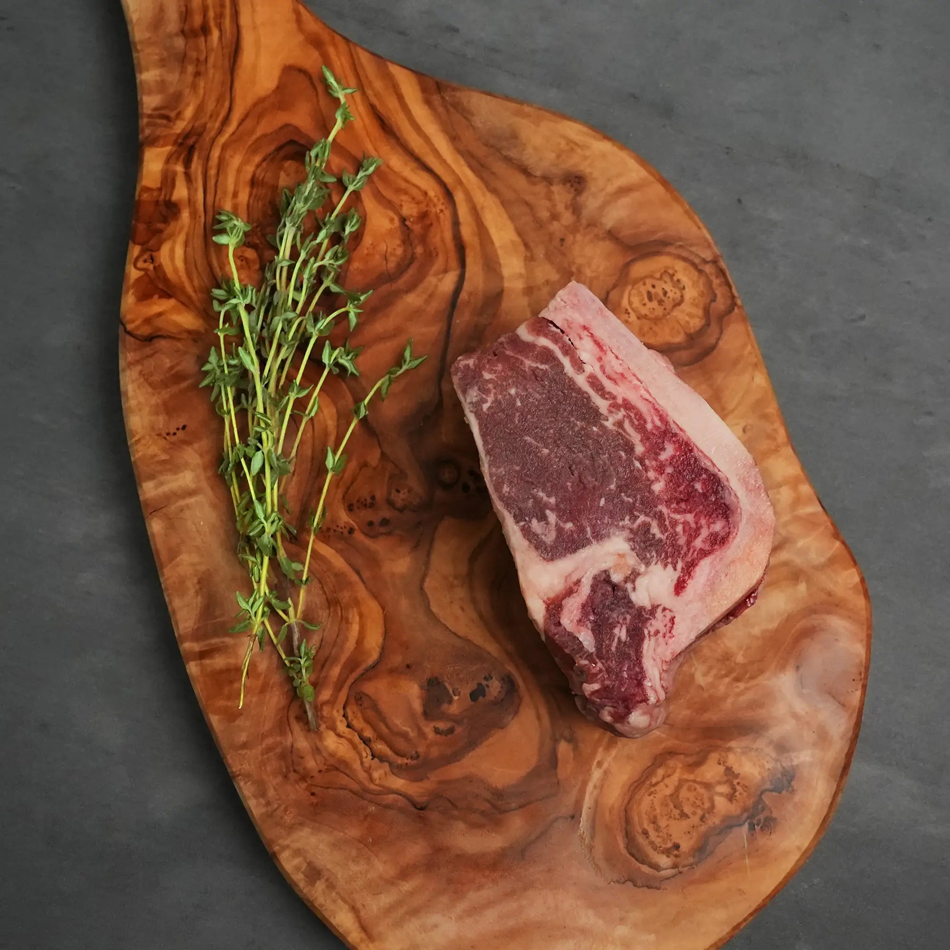 Choice Bone-In Filet Mignon on olive-wood board with fresh thyme, showcasing its marbling and bone for tender, juicy steakhouse-quality cooking.