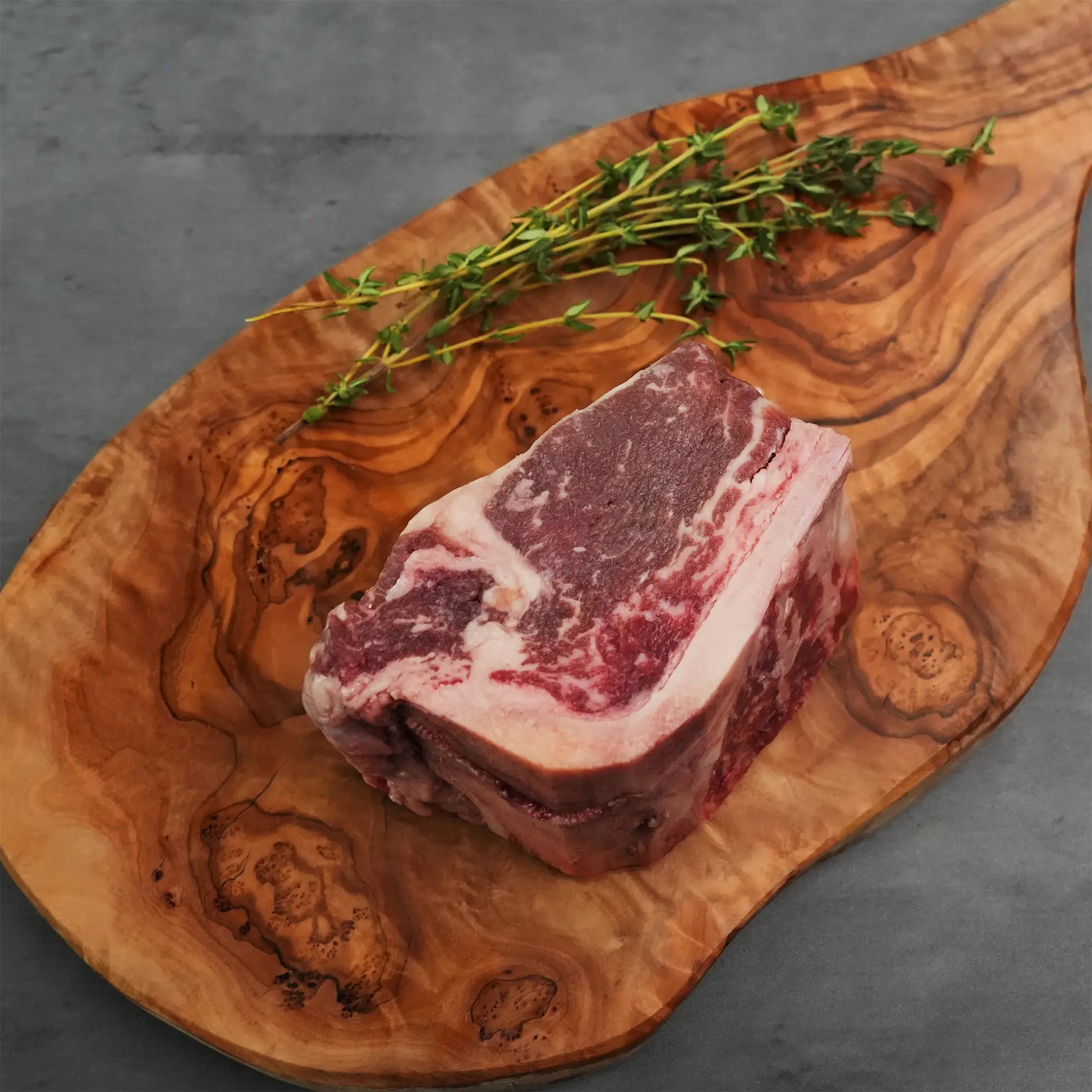 Choice Bone-In Filet Mignon on rustic board, well-marbled and wet-aged, accompanied by thyme sprigs, promising steakhouse quality with tender, juicy texture.