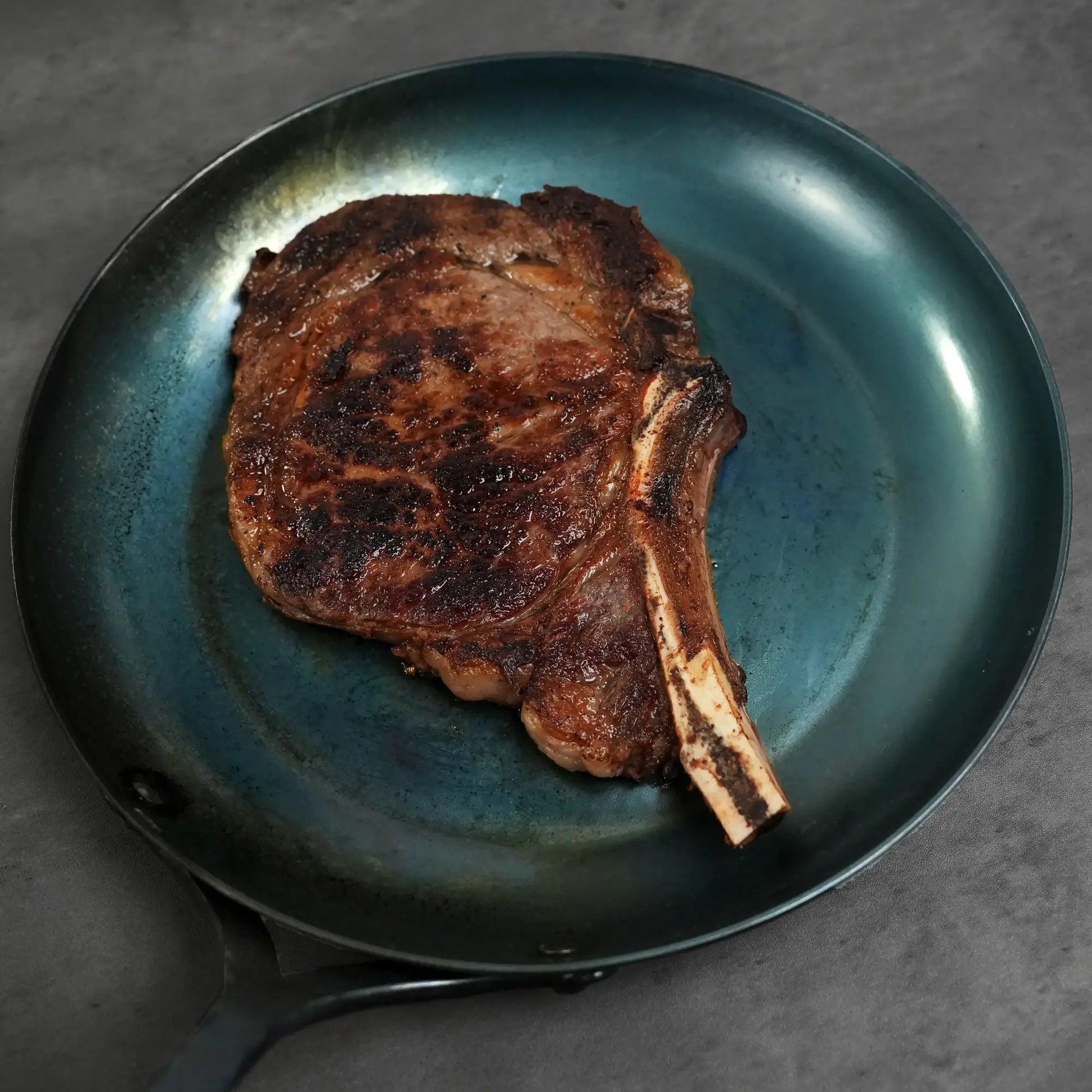 Prime Split-Bone Ribeye Steak seared to perfection, resting in a shallow teal pan, highlighting its hand-cut thickness and rich flavor for easy preparation.