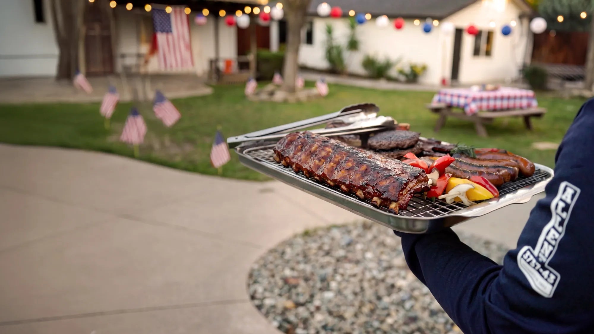 Person holding a tray of barbecue ribs and vegetables outdoors with a house and garden in the background.