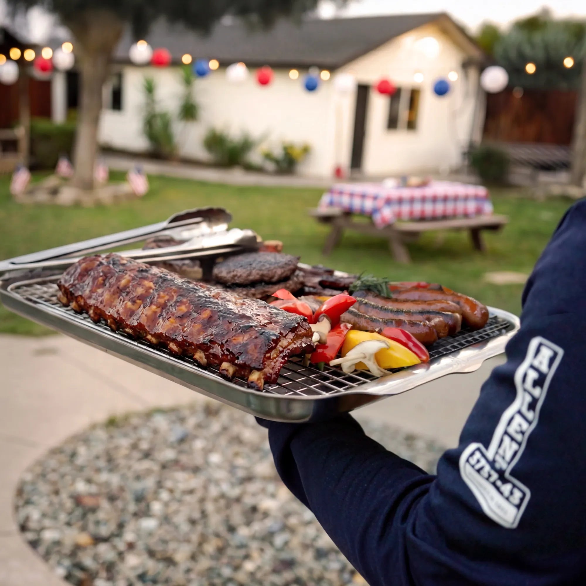 Person holding a tray of grilled ribs and vegetables in a backyard setting with a picnic table and string lights.