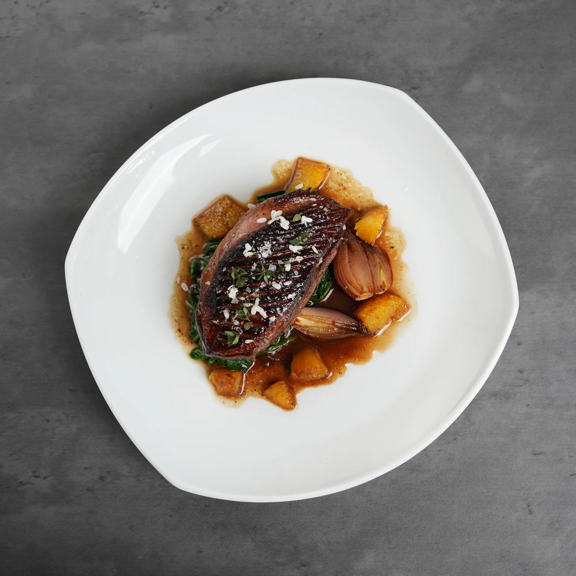 Skin On Magret Duck Breast, seared and glazed, rests on wilted greens with caramelized shallots and roasted squash cubes, garnished with herbs and coarse salt.