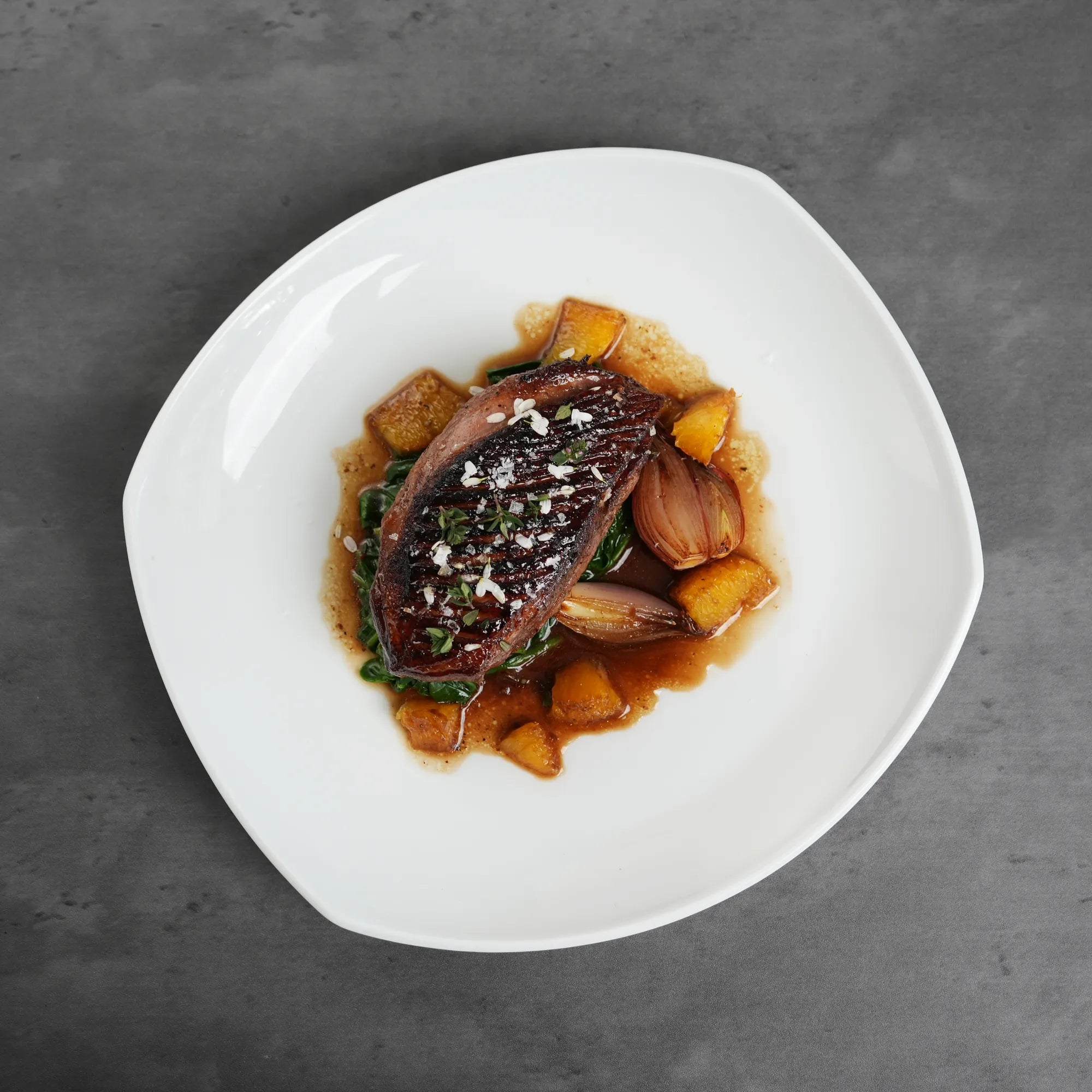 Skin On Magret Duck Breast, seared and glazed, rests on wilted greens with caramelized shallots and roasted squash cubes, garnished with herbs and coarse salt.