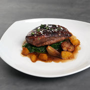 Skin On Magret Duck Breast plated with flaky sea salt, fresh herbs, wilted greens, roasted shallots, and orange segments in a rich pan sauce.