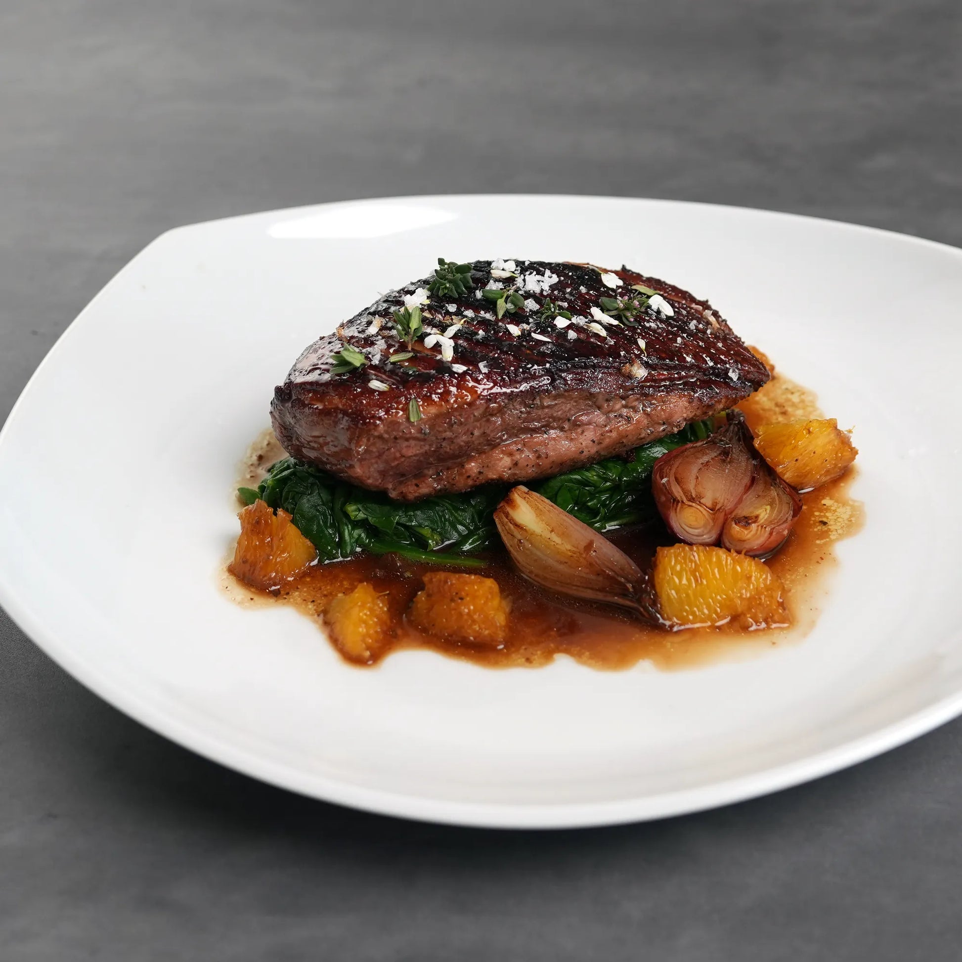 Skin On Magret Duck Breast plated with flaky sea salt, fresh herbs, wilted greens, roasted shallots, and orange segments in a rich pan sauce.