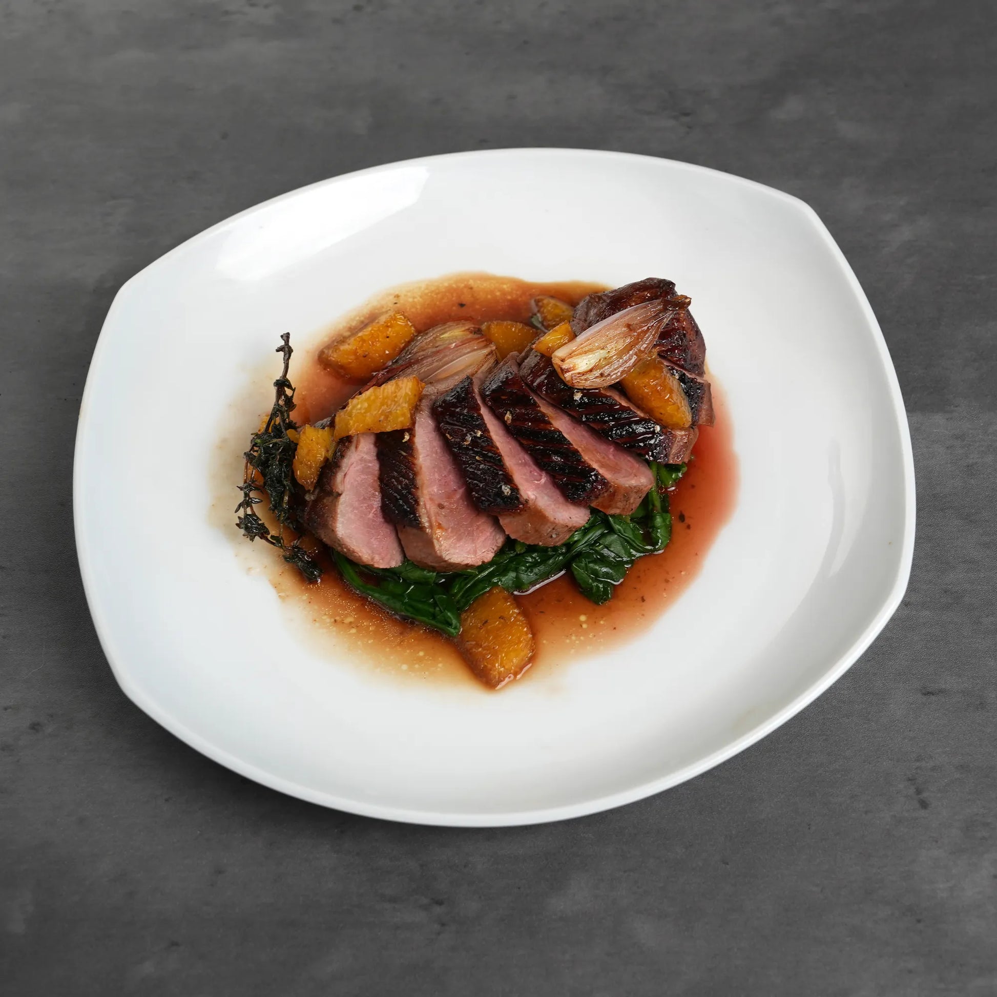 Skin On Magret Duck Breast, medium-rare slices on wilted greens, topped with orange sauce, segments, caramelized shallots, and thyme on a white plate.