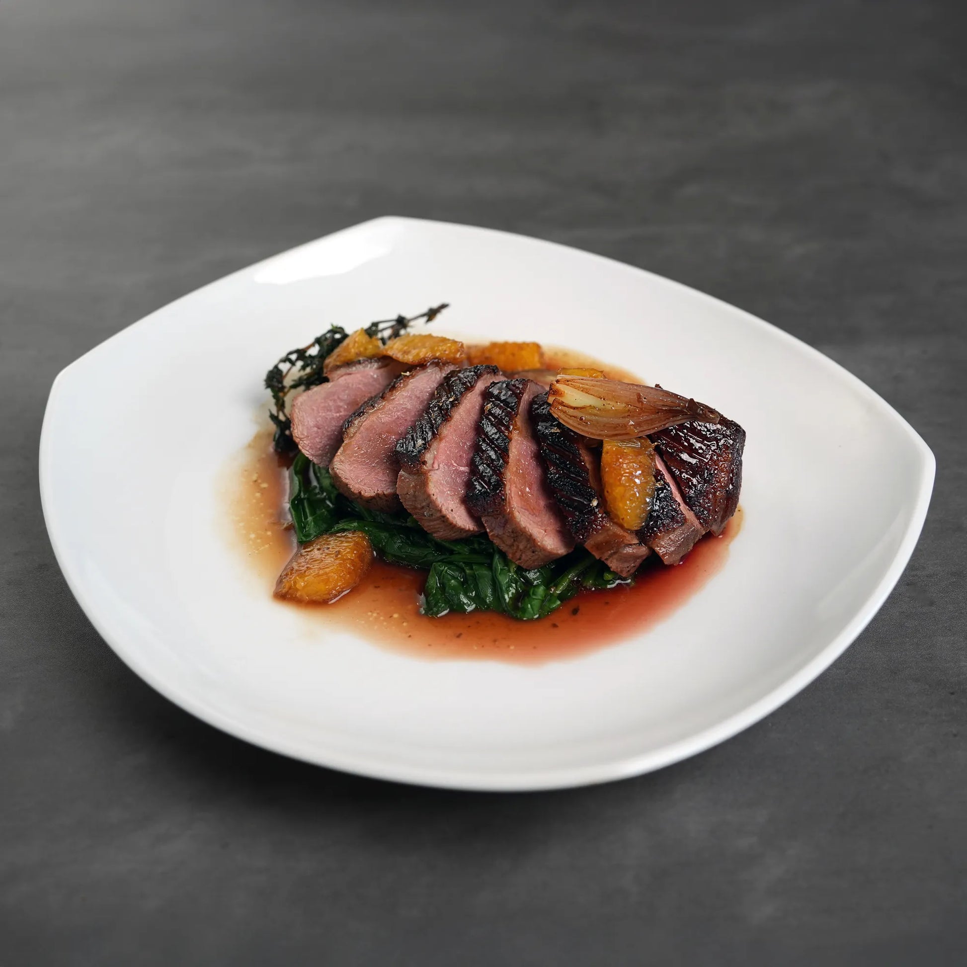 Skin On Magret Duck Breast slices on wilted greens with citrus, drizzled with pan sauce, highlighting the entrée's richness and suitability for rare to medium-rare cooking.