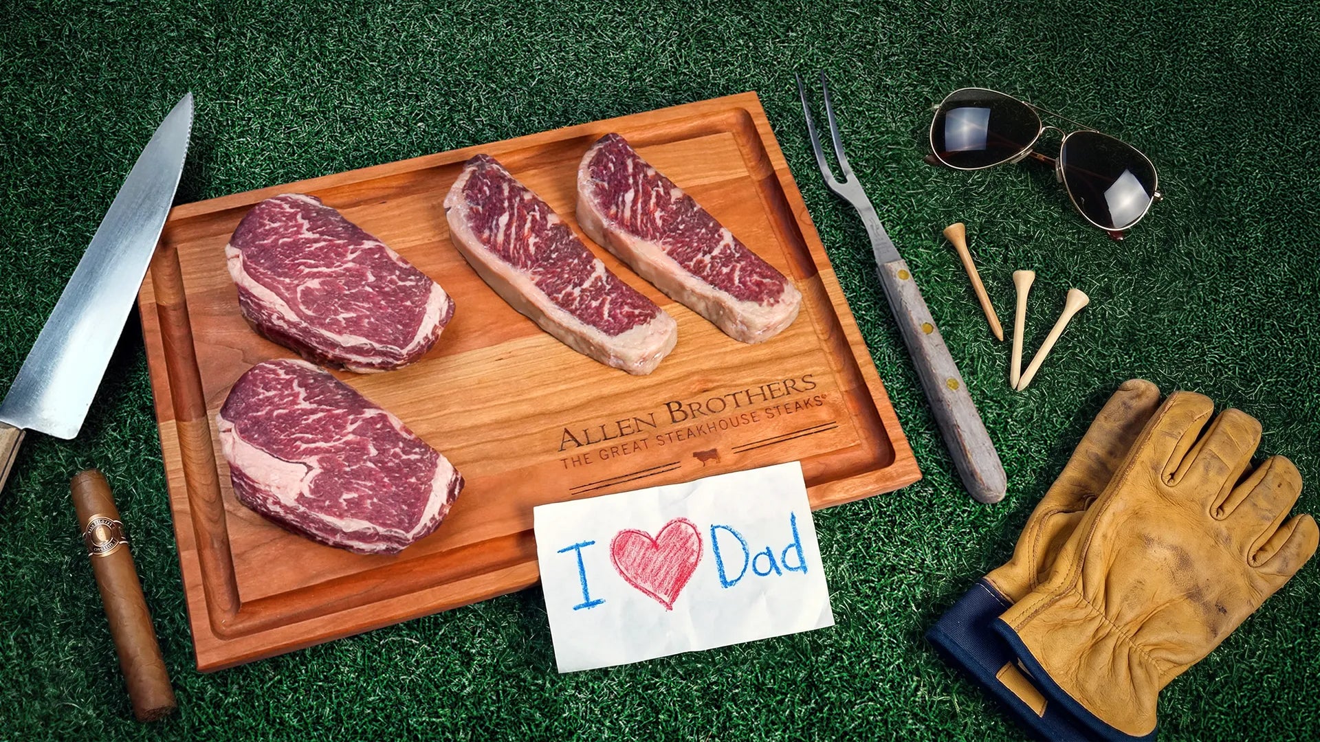 Steaks on a cutting board with a 'I ❤️ Dad' card, knife, fork, sunglasses, and gloves on a grassy background.
