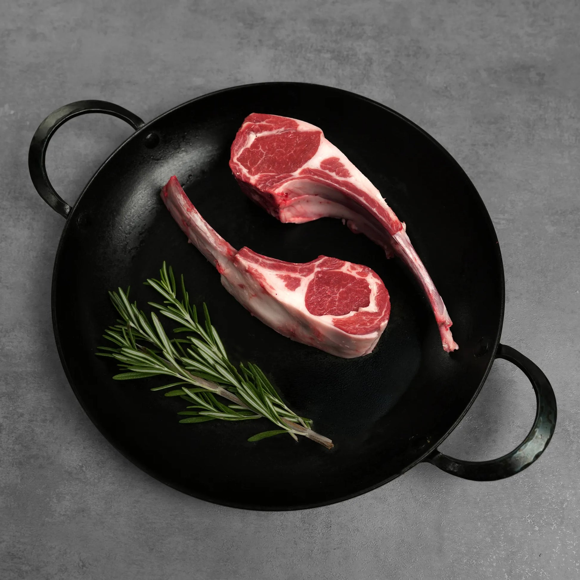 Double Cut Halal Australian Lamb Chop, featuring two raw lollipop-style chops in a black cast-iron pan with fresh rosemary sprigs, ready for cooking.