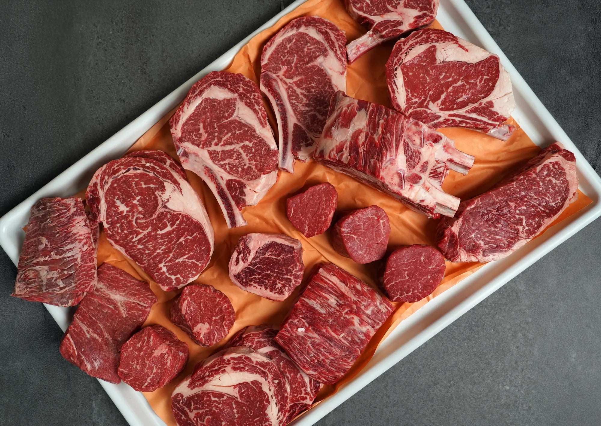 Assorted cuts of raw red meat on a white tray with a dark background