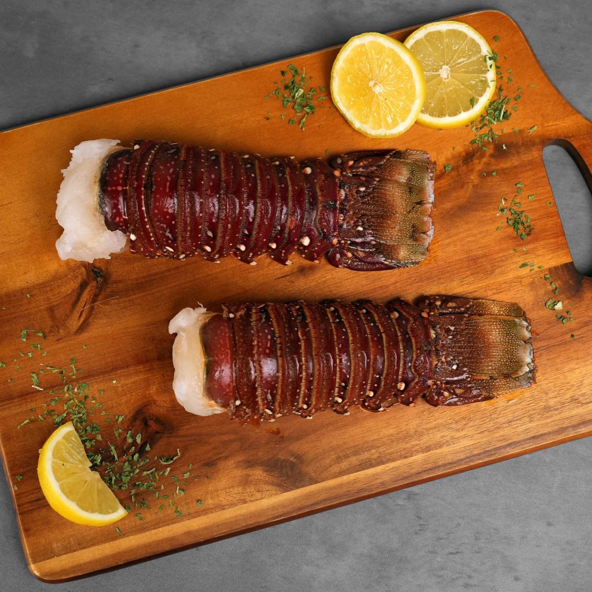Surf & Turf XL Combo: Two spiny-lobster tails on a wooden board, meat exposed, garnished with lemon slices and chopped herbs, showcasing premium seafood quality.