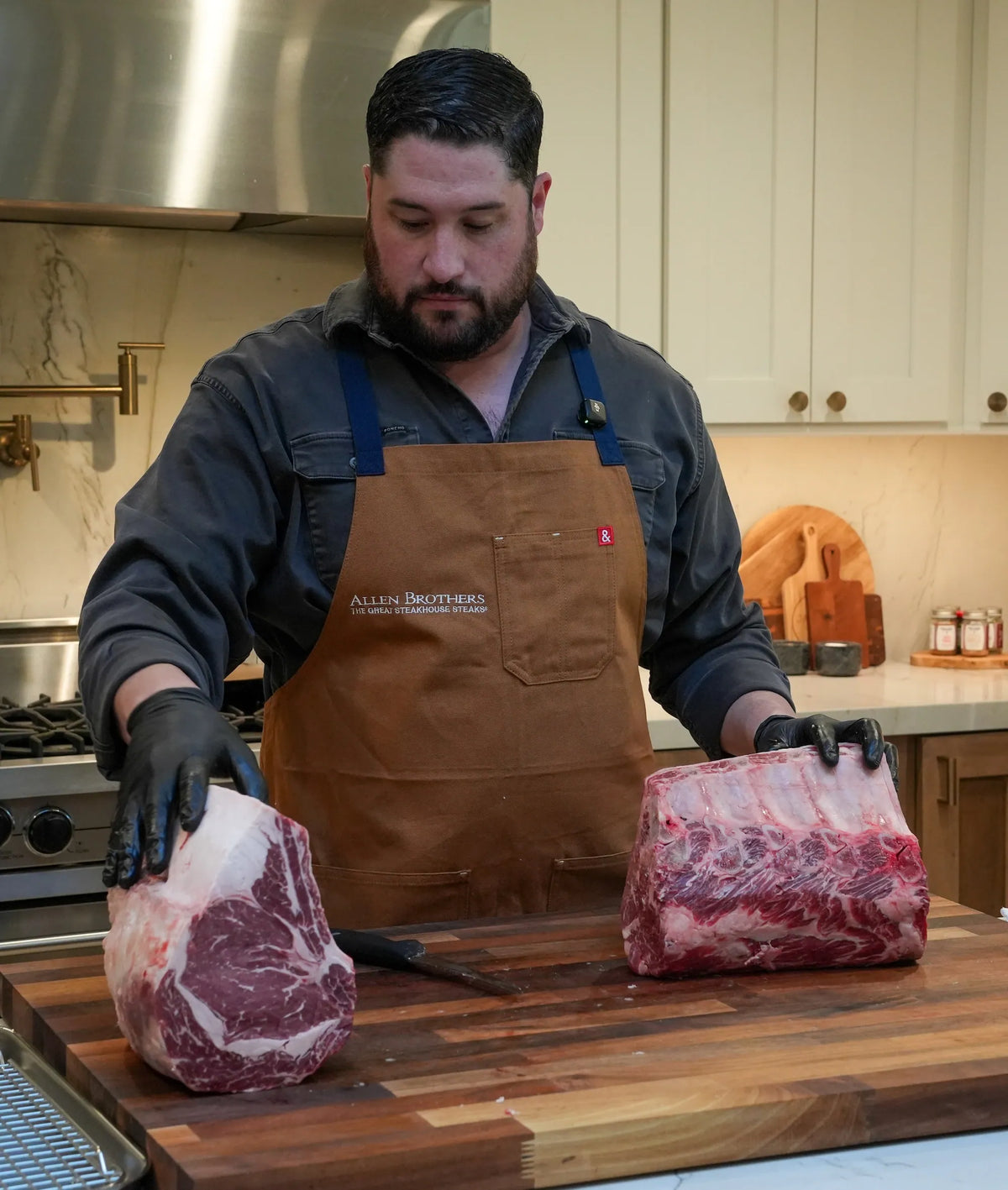 A man wearing a brown leather apron and black gloves stands behind a wooden cutting board in a kitchen, holding two large raw beef brisket racks.