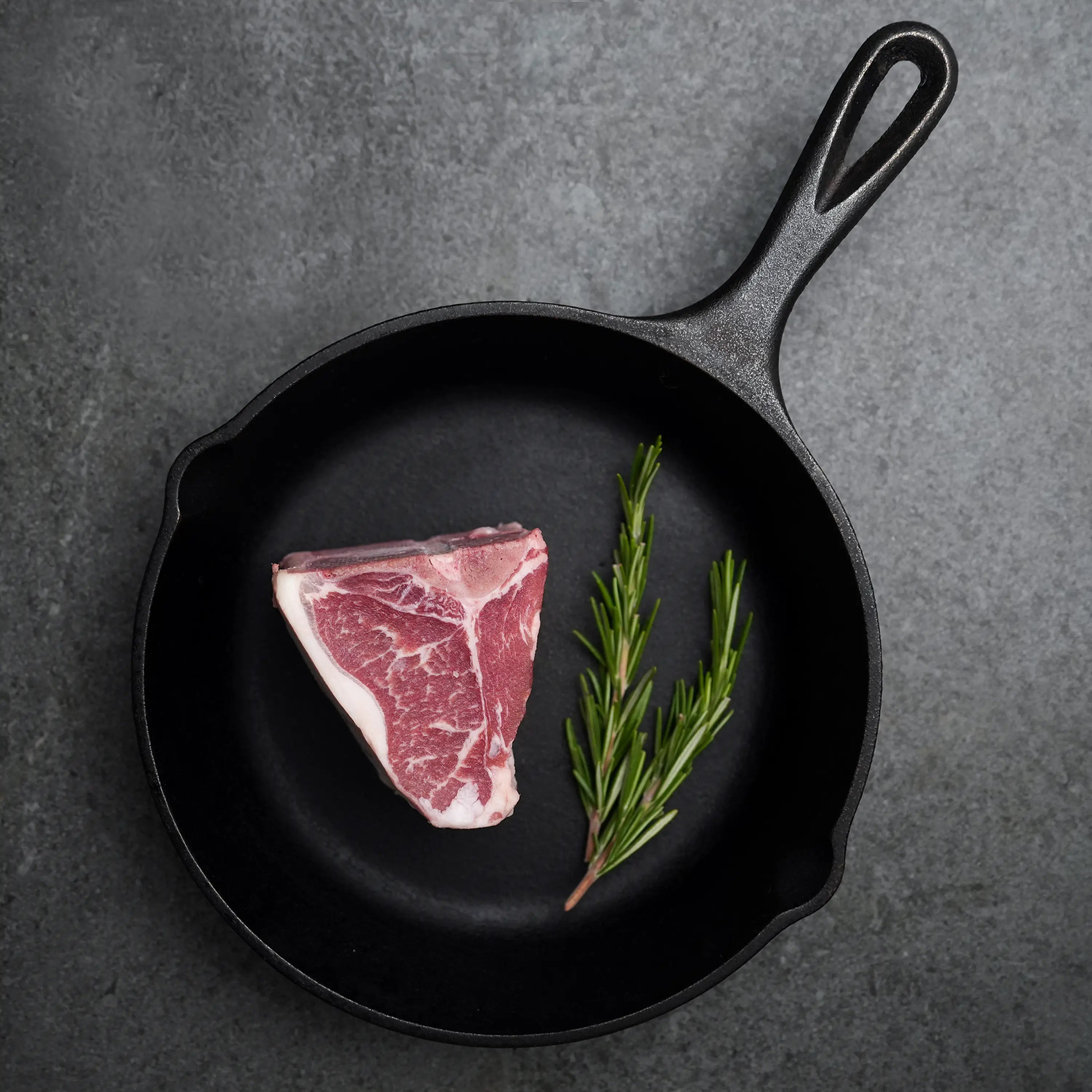 Domestic All Natural Lamb Loin T-Bone Chops in a cast-iron skillet with fresh rosemary, ready for searing.