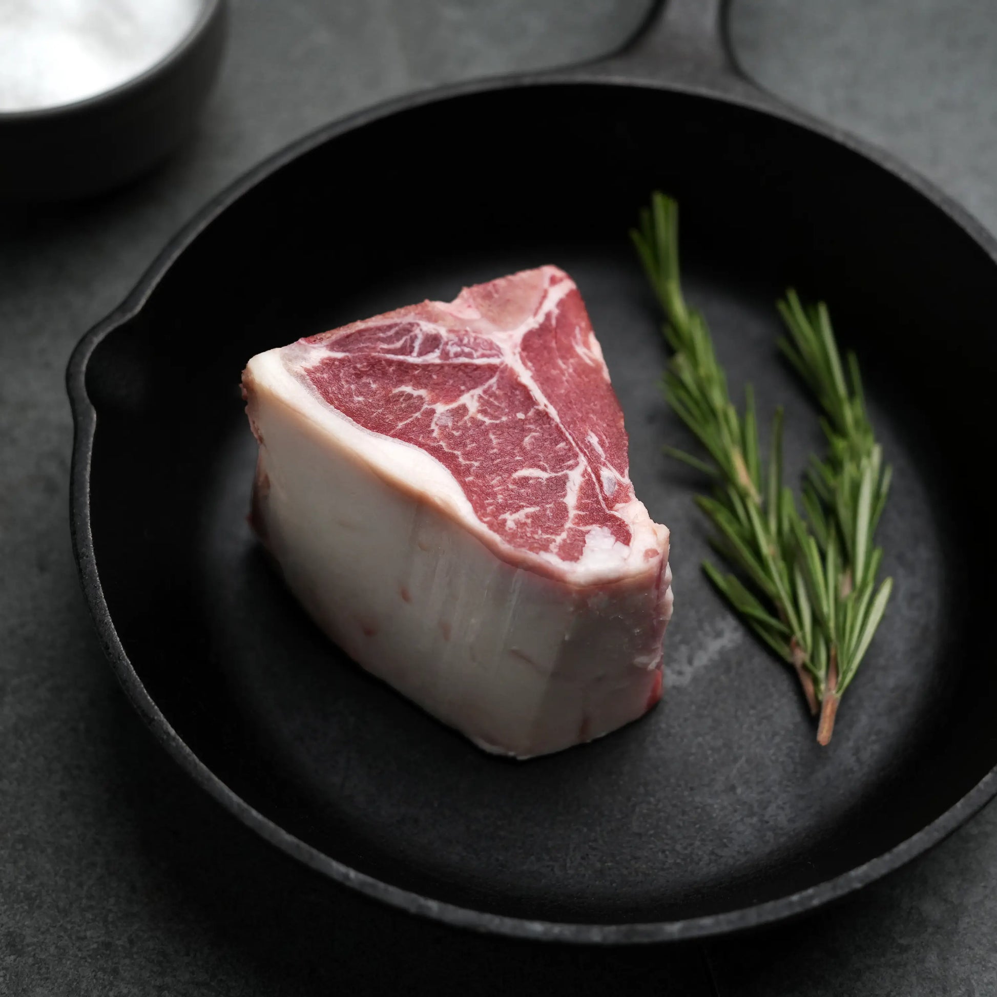 Domestic All Natural Lamb Loin T-Bone Chops in a cast-iron skillet with rosemary sprigs, highlighting their marbled texture and suitability for simple, flavorful cooking.