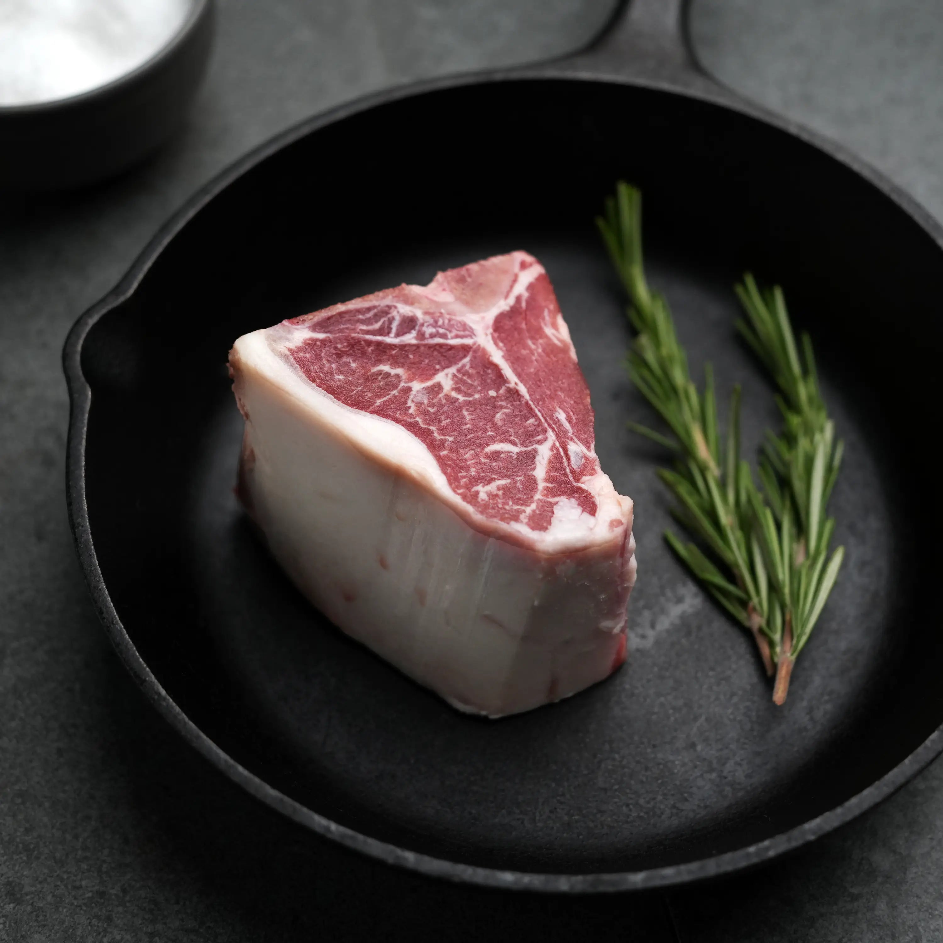 Domestic All Natural Lamb Loin T-Bone Chops in a cast-iron skillet with rosemary sprigs, highlighting their marbled texture and suitability for simple, flavorful cooking.