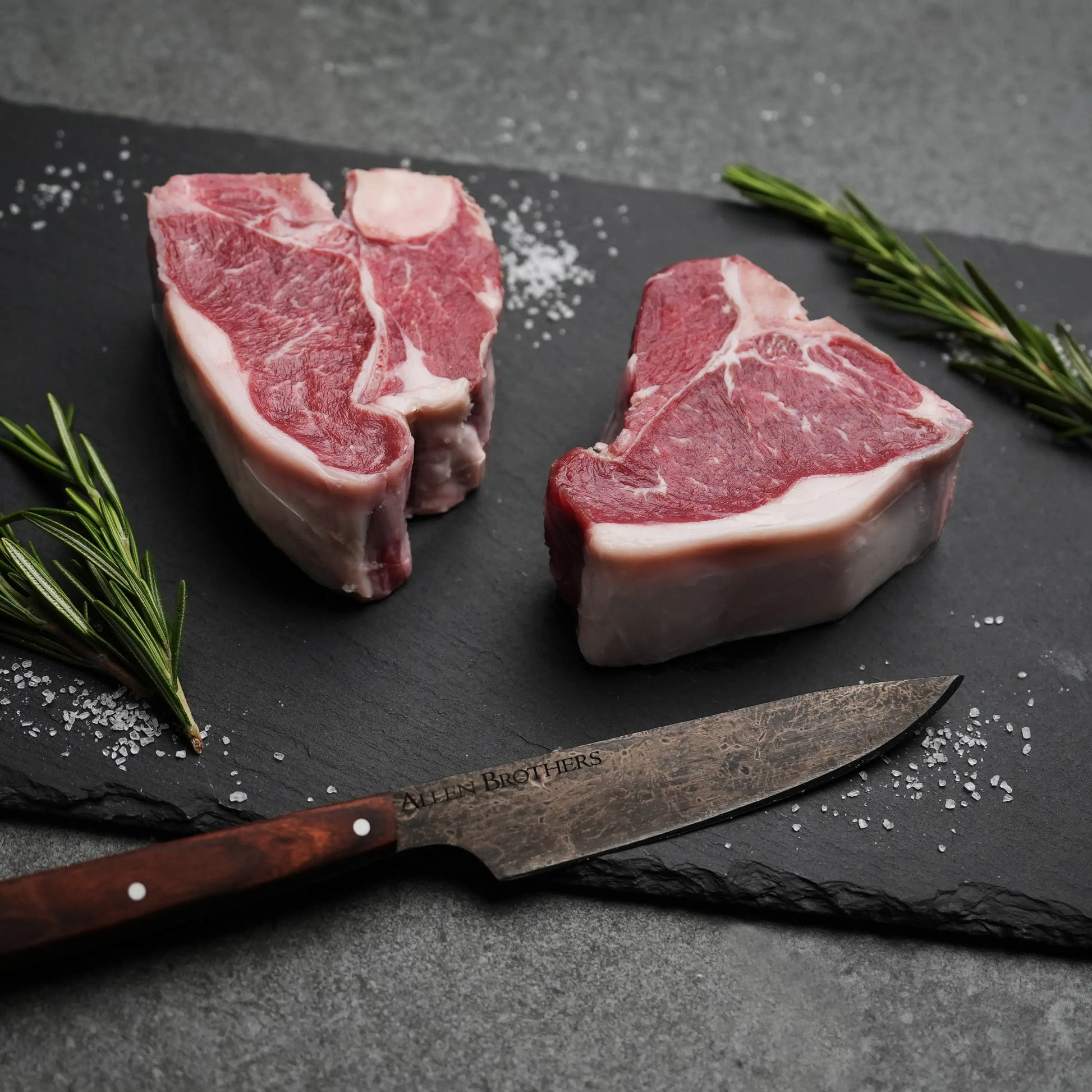 Domestic All Natural Lamb Loin T-Bone Chops, uncooked on slate, with rosemary sprigs, coarse salt flakes, and a butcher’s knife beside.