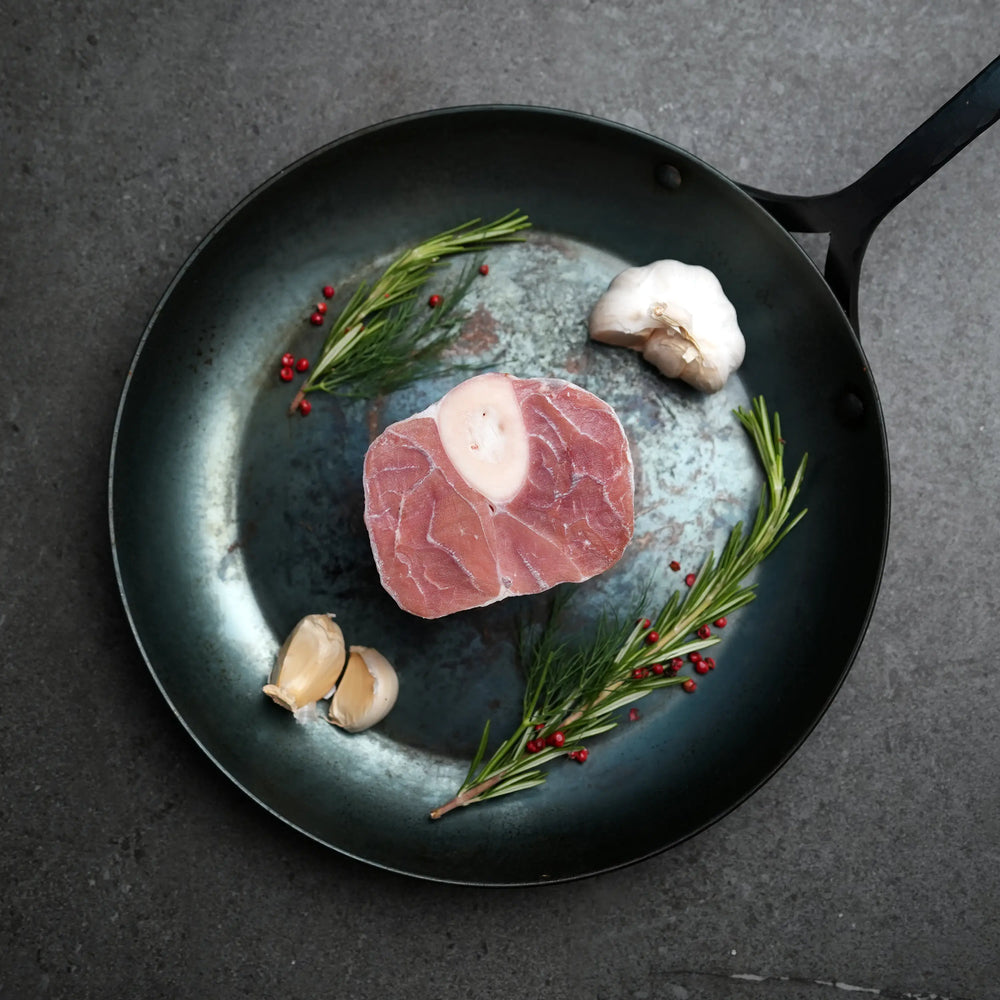 Milk Fed Dutch Veal Osso Buco Shank in skillet with garlic, rosemary, and red peppercorns, showcasing its raw, bone-in cut ready for braising.